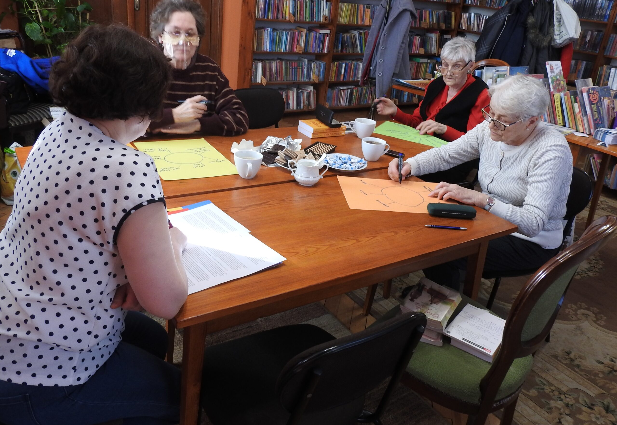 Moderatorka oraz członkinie Dyskusyjnego Klubu Książki dla dorosłych podczas oraz moderatorka podczas spotkania w bibliotece. Klubowiczki na kolorowych kartkach wpisują naokoło okręgu elementy z otaczającej ich przestrzeni zewnętrznej.