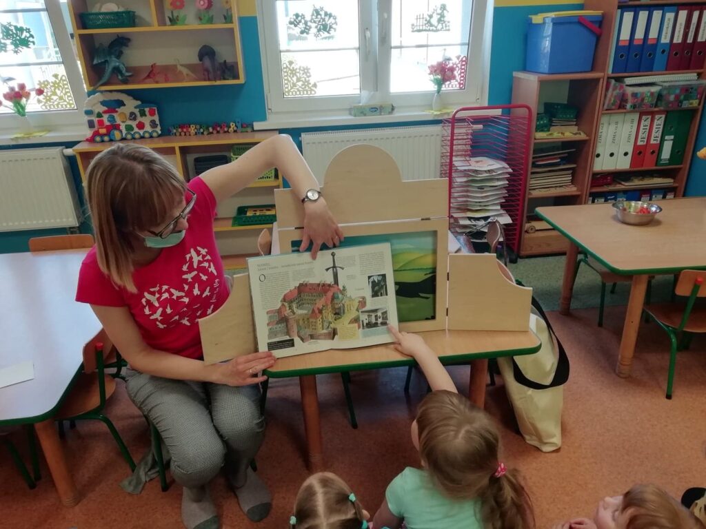 Kobieta siedzi na małym krzesełku i pokazuje ilustracje w książce siedzącym przed nią dzieciom.