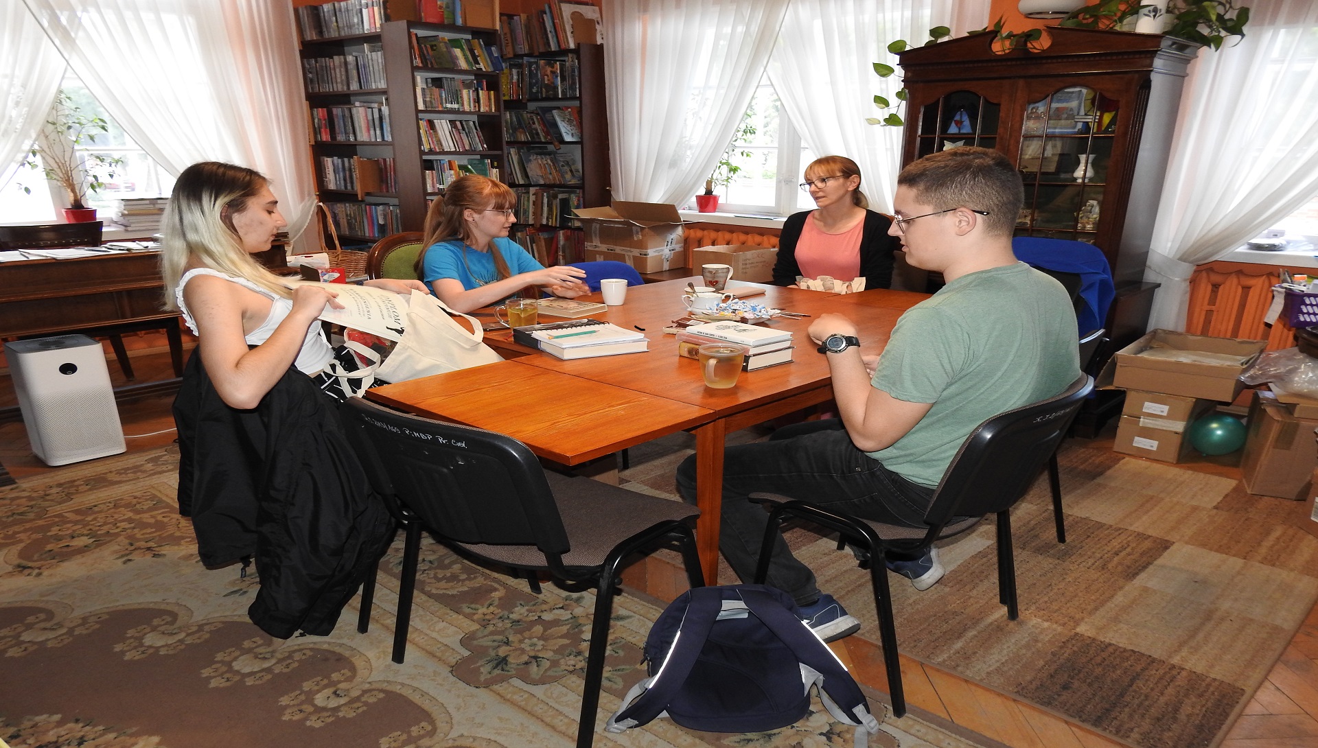 Cztery osoby siedzą przy stole wewnątrz pomieszczenia. Za nimi regały z książkami.