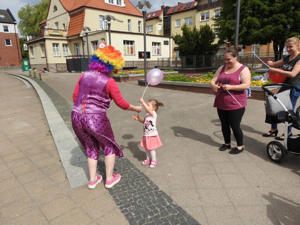 Postać w kolorowym stroju i peruce wręcza balonika małej dziewczynce. Zza dzieckiem stoi kobieta.