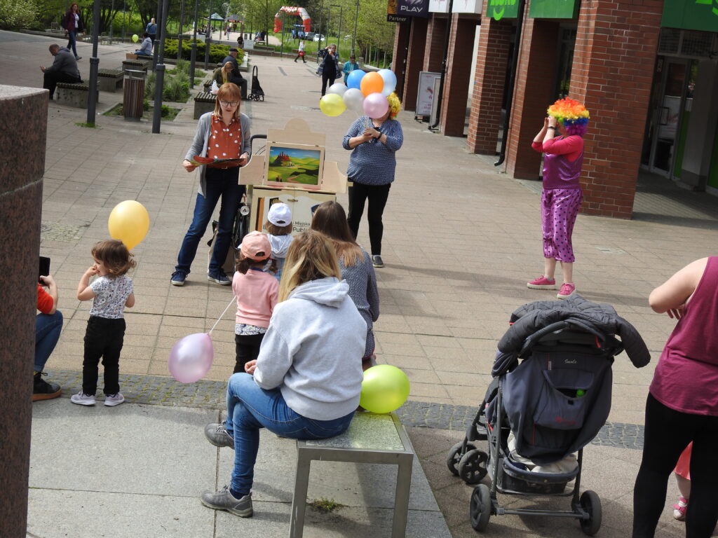 Grupa dorosłych i dzieci zwrócona w stronę 3 osób, z których jedna stoi obok drewnianej skrzynki z kolorowymi obrazkami, czyta. Obok osoba w balonami, a z jej boku postać w stroju clowna.