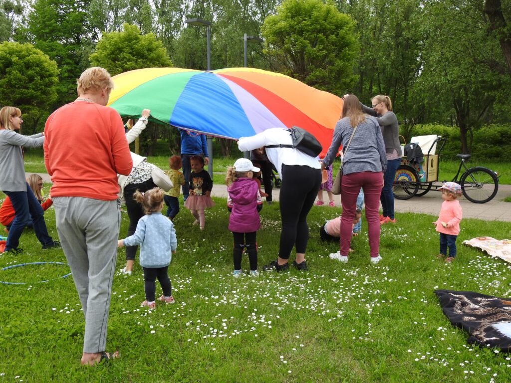 Grupa dorosłych oraz dzieci na świeżym powietrzu podczas zabawy z różnokolorową chustą.