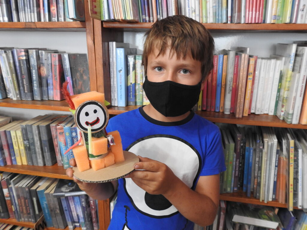 Dziecko w masce, w tle regały z książkami, w ręce trzyma figurkę wykonaną z różnych materiałów. 