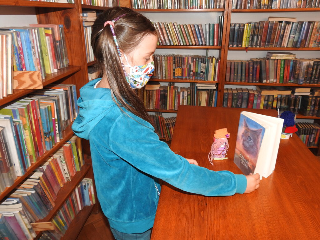 Dziewczynka patrzy na książkę ustawiona na stole, obok którek stoi kolorowa figurka. W tle regały z książkami.