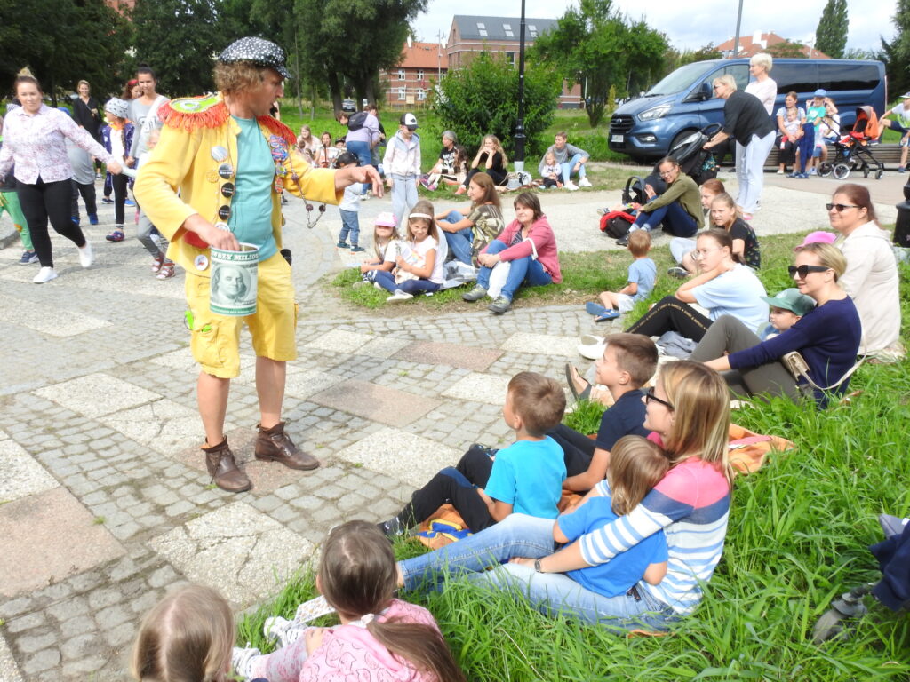 Postać w żółtym stroju kieruje mikrofon w stronę osób dorosłych i dzieci, które siedzą na kocu.