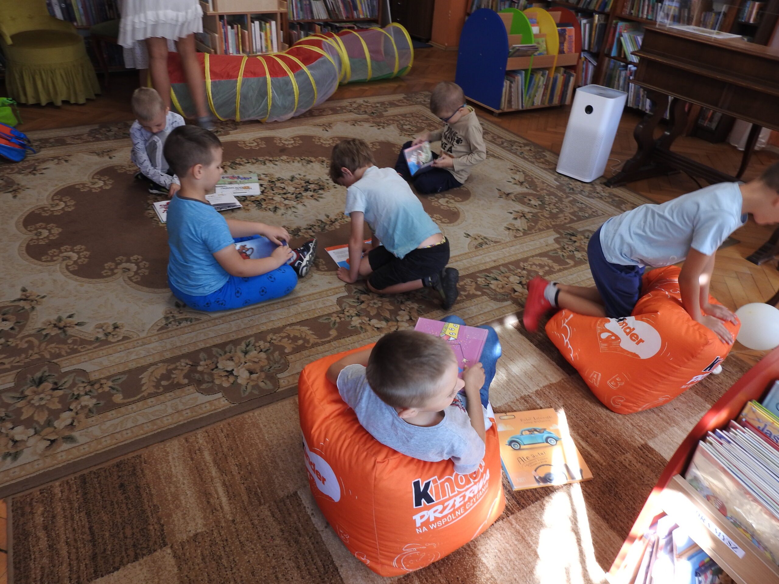 Grupa dzieci wewnątrz pomieszczenia, w tle regały z książkami, napis: droga książki do autora do czytelnika, Powiatowa i Miejska Biblioteka Publiczna w Pruszczu Gdańskim
