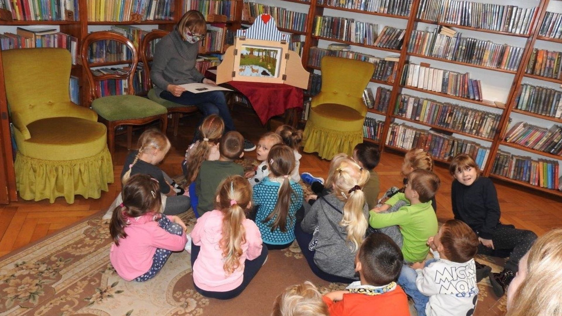 Jedna osoba siedzi przodem do grupy dzieci. Obok niej na stole drewniane otwarte pudełko z kolorową planszą w środku. W tle regały z książkami.