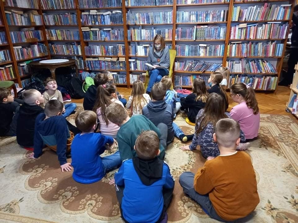 Grupa dzieci siedzących na dywanie w pomieszczeniu z książkami. Przed nimi na krześle siedzi osoba dorosła z książka w ręku.