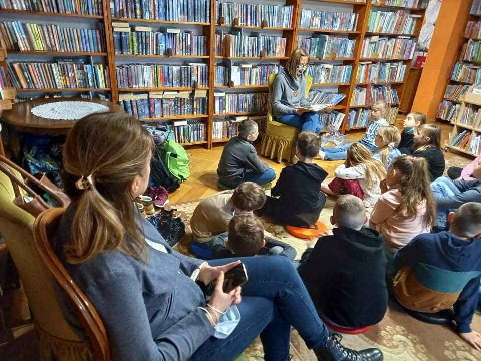 Grupa dzieci siedzących na dywanie w pomieszczeniu z książkami. Przed nimi na krześle siedzi osoba dorosła z książka w ręku.