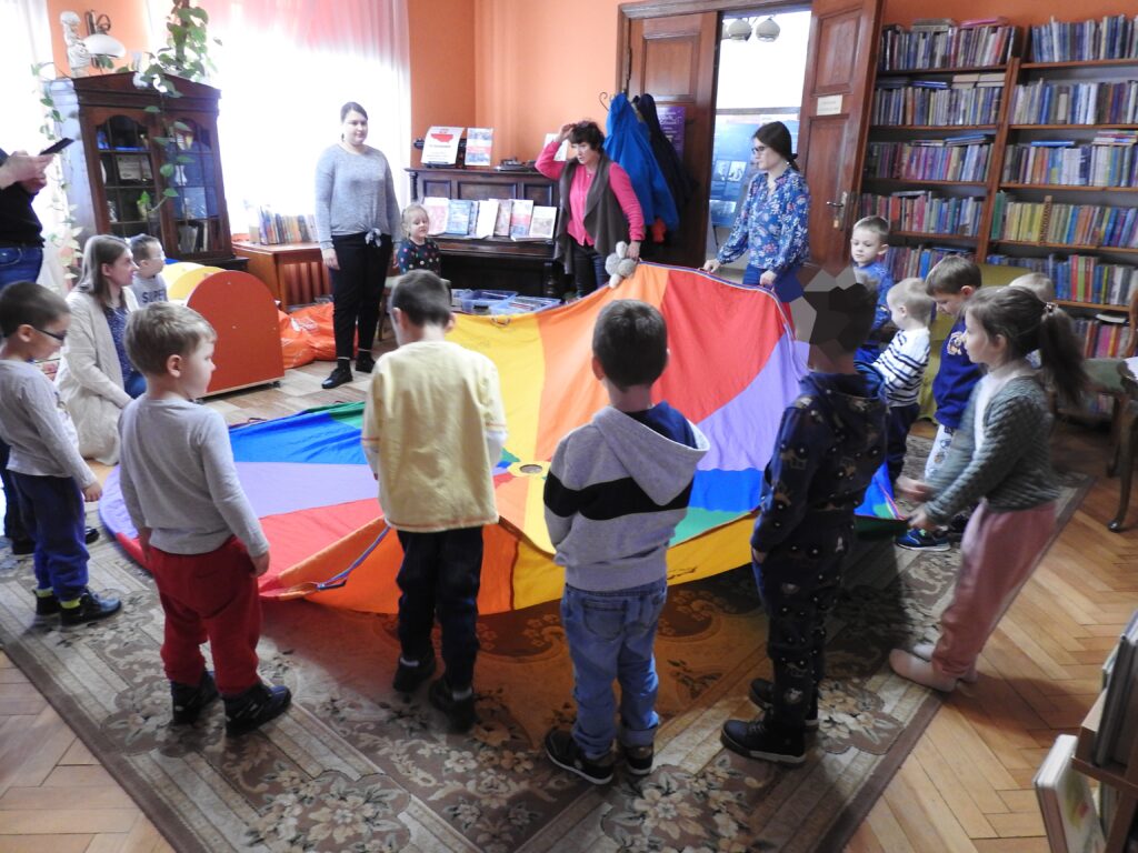 Zdjęcie wewnątrz pomieszczenia. Kilkoro dorosłych oraz dzieci stoi w okregu i trzyma kolorowa płachtę.