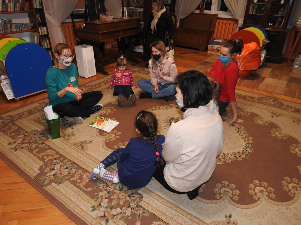 cztery osoby dorosłe oraz troje dzieci na siedzi na dywanie w pomieszczeniu z książkami.
