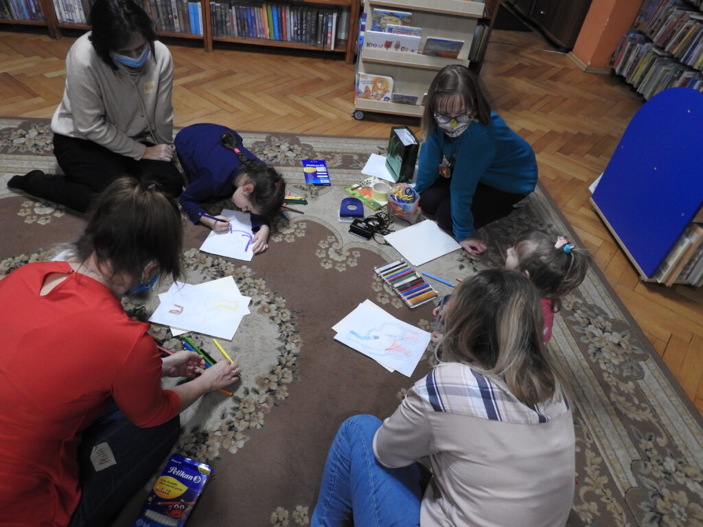 cztery osoby dorosłe i dwójka dzieci na dywanie, w pomieszczeniu z ksiązkami. Przed nimi leżą zarysowane kartki i kredki.