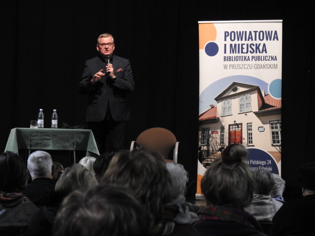 Mężczyzna na scenie z mikrofonem w ręku. Przed nim siedzi kilkanaście osób. Obok baner z napisem: Powiatowa i Miejska Biblioteka Publiczna w Pruszczu Gdańskim.