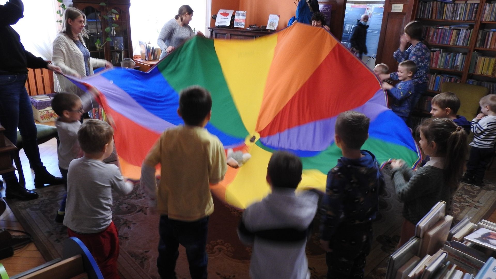 Zdjęcie wewnątrz pomieszczenia. Kilkoro dorosłych oraz dzieci stoi w okregu i trzyma kolorowa płachtę.