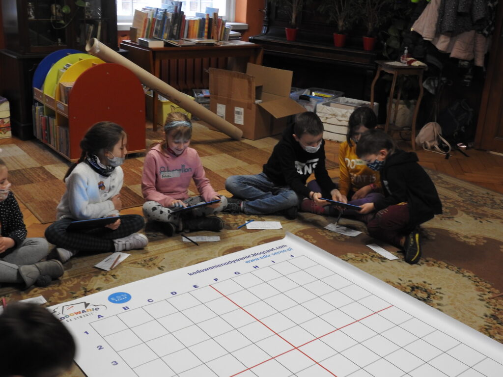 Na dywanie leży mata z napisem:kodowanie na dywanie,  wokół niej kilkoro dzieci.ty nich tablety oraz robot.