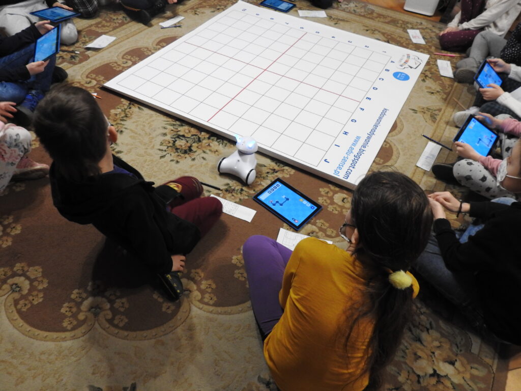 na dywanie mata z napisem: kodowanie na dywanie, wokół nioej siedzą osoby, które trzymają lub leżą obok tablety nich tablety oraz robot.