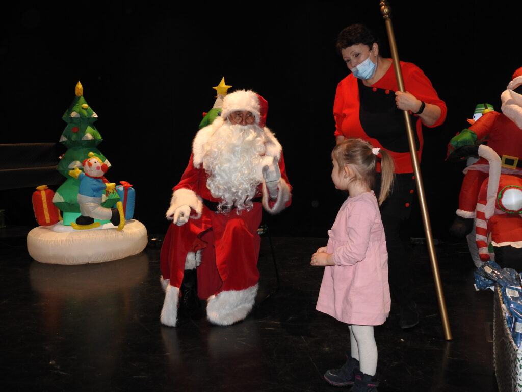 Święty Mikołaj i kobieta, obok nich mała dziewczynka w rózowej sukience. W tle dmuchana choinka.