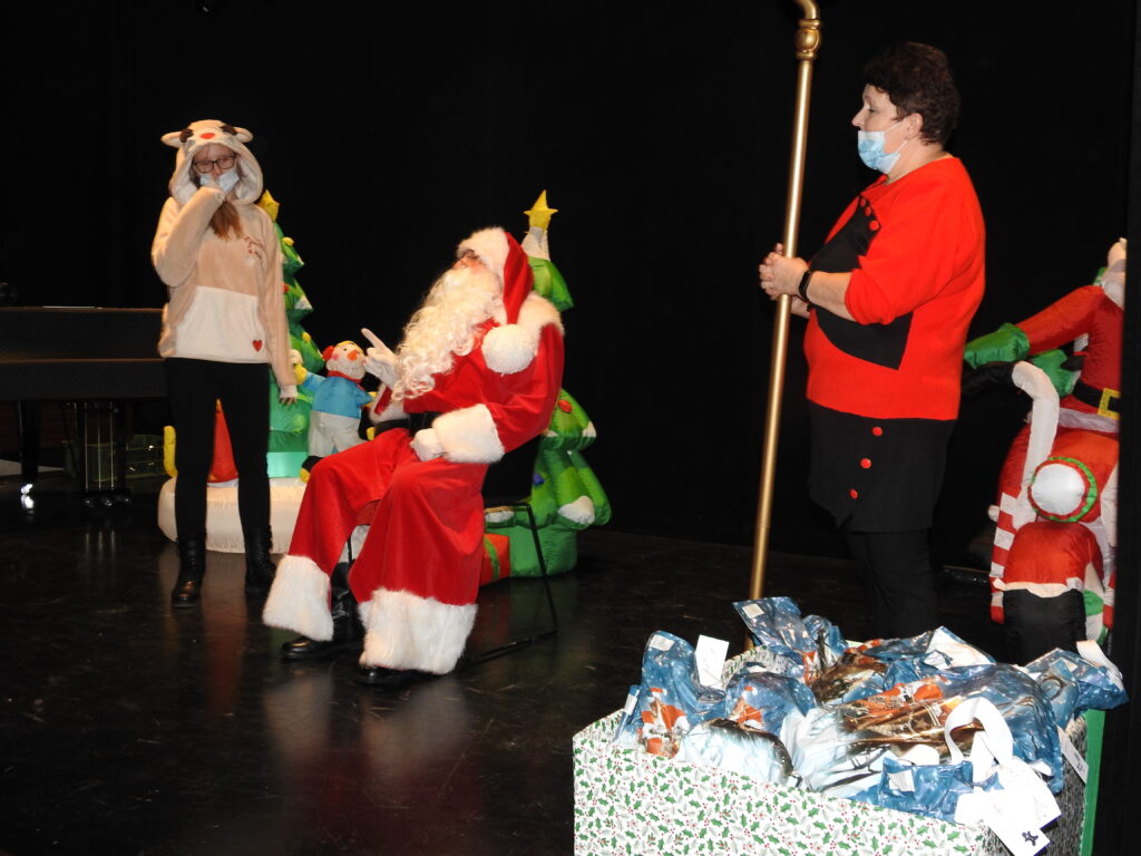 Święty Mikołaj zwrócony w stronęn dziecka oraz postać stojąca obok z laską Mikołaja. Z przodu karton z prezentami. Za nimi dmuchane ozdoby: choinka oraz Mikołaj na motorze.