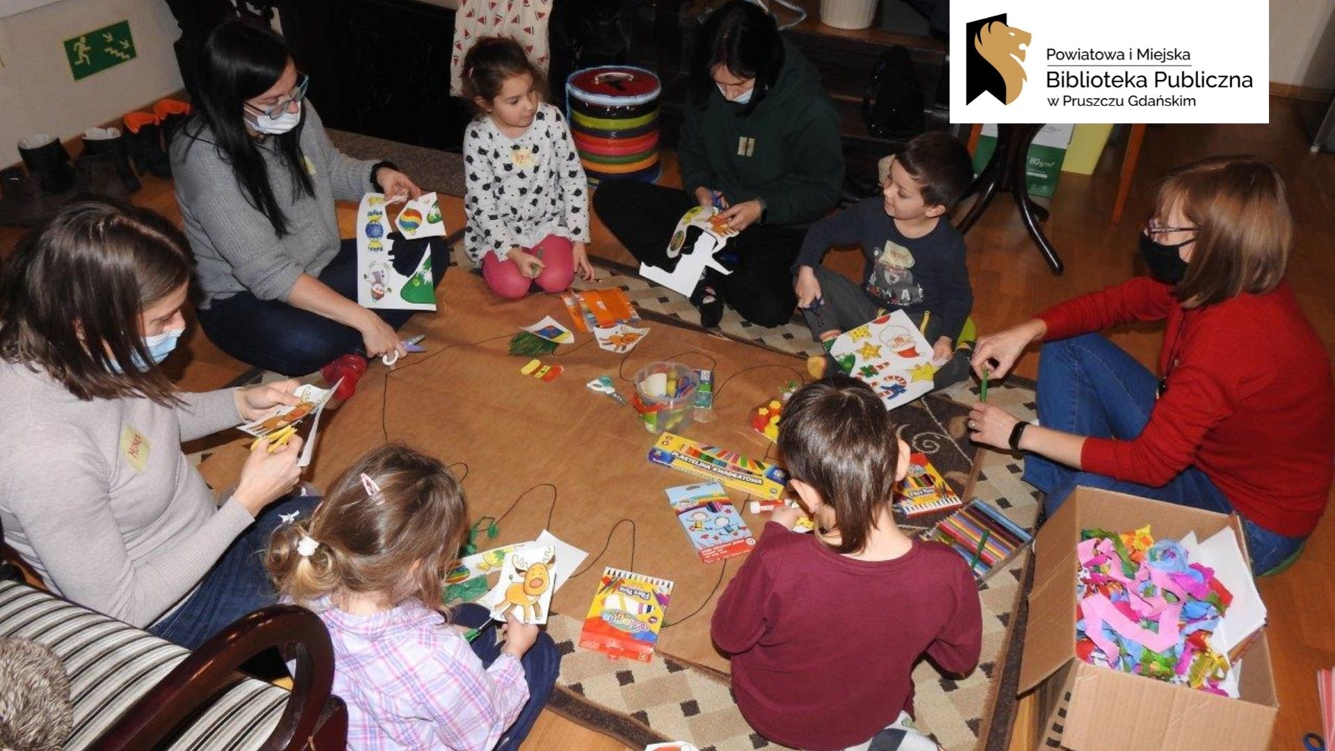 Czworo dorosłych oraz czwórka dzieci siedzi na dywanie i wycinają kolorowe: bałwanki, gwiazdki i bombki. Dzieci trzymają w rzekach kleje, obok leżą mazaki, fragmenty kolorowej bibuły.