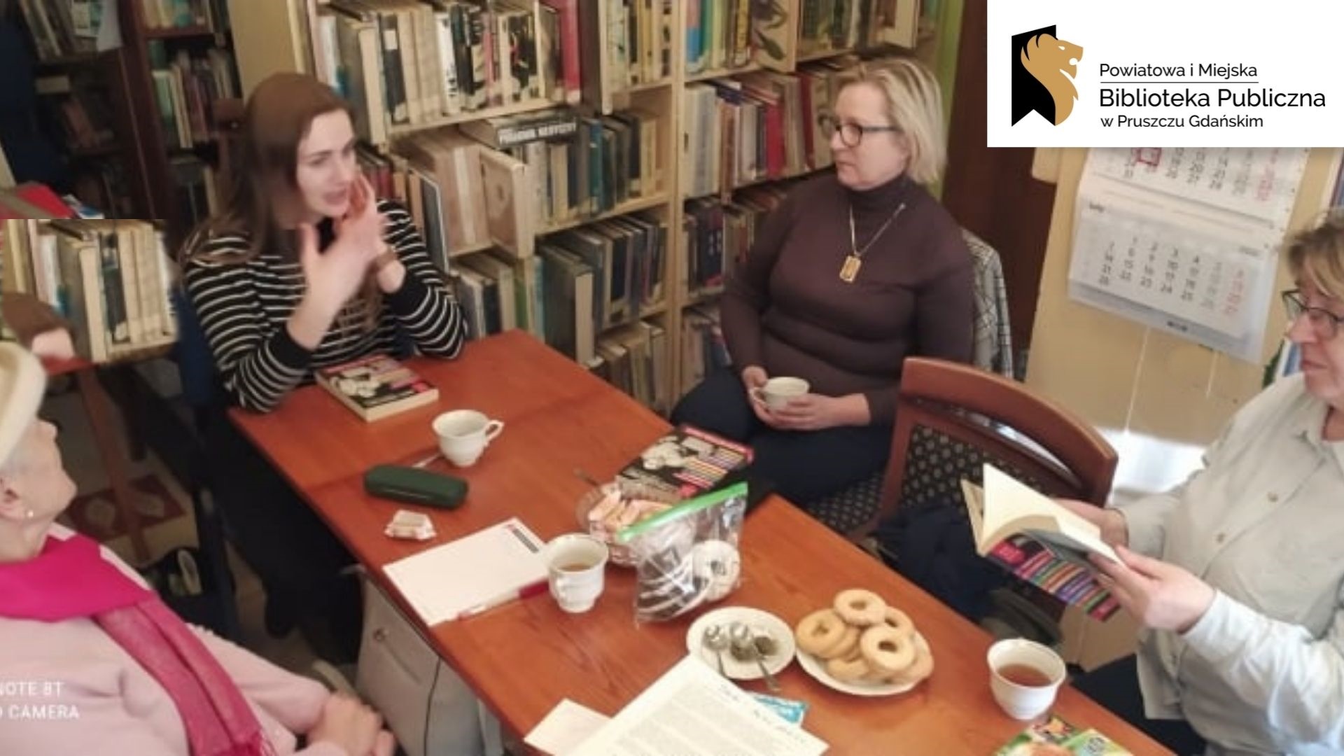 Cztery osoby siedzą przy stole. W tle regały z książkami. Logotyp oraz napis: Powiatowa i Miejska Biblioteka Publiczna w Pruszczu Gdańskim.