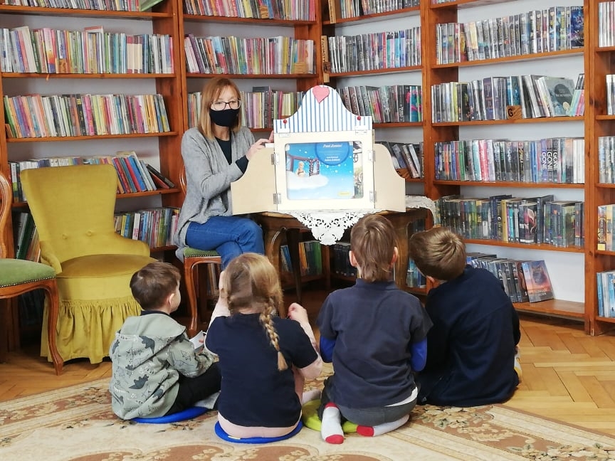 Czworo dzieci siedzi na dywanie, zwrócona w stronę osoby siedzącej na krześle, przed którą stoi drewniana skrzynka z obrazkiem w środku. Za osobą książki na regałach.
