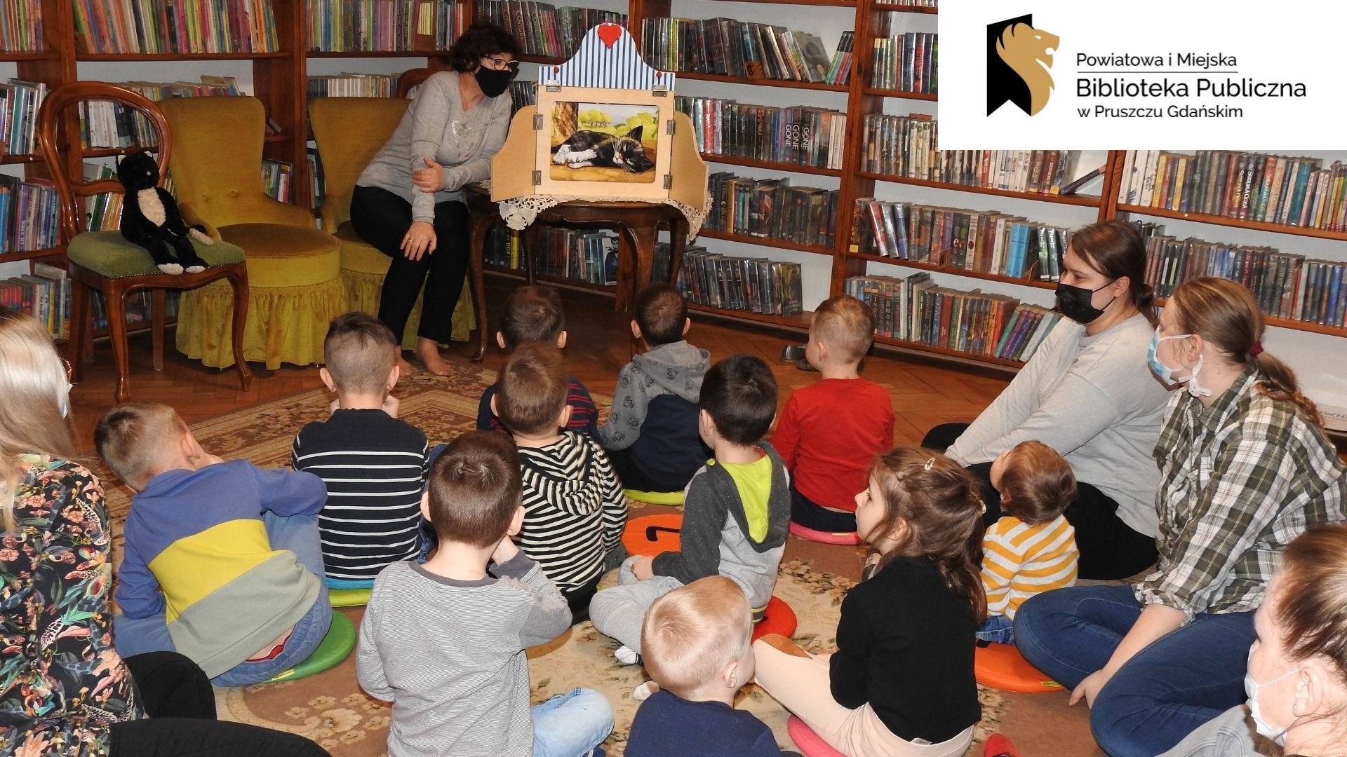 Dzieci siedzą na dywanie zwrócone w stronę osoby dorosłej, obok której stoi drewniana skrzynka z ilustracją w środku. Za nimi regały z książkami