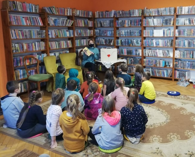 Grupa dzieci siedzi na dywanie, zwrócona w stronę osoby siedzącej na krześle, przed którą stoi drewniana skrzynka. Za osobą książki na regałach.