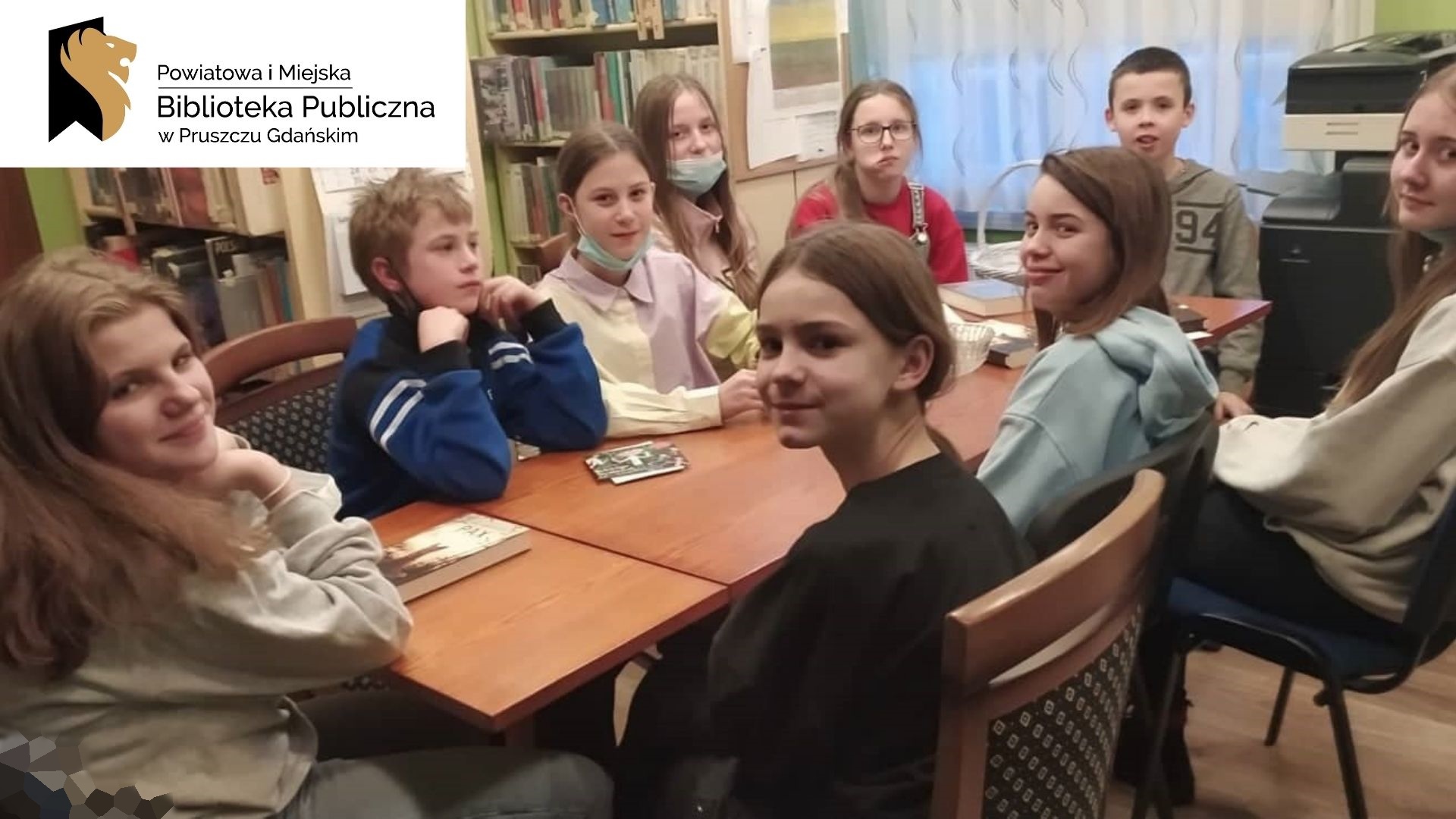 Dziewięcioro dzieci siedzi przy stole z twarzami zwróconymi w stronę osoby fotografującej. Logotoyp z napisem Powiatowa i Miejska Biblioteka Publiczna w Pruszczu Gdańskim.