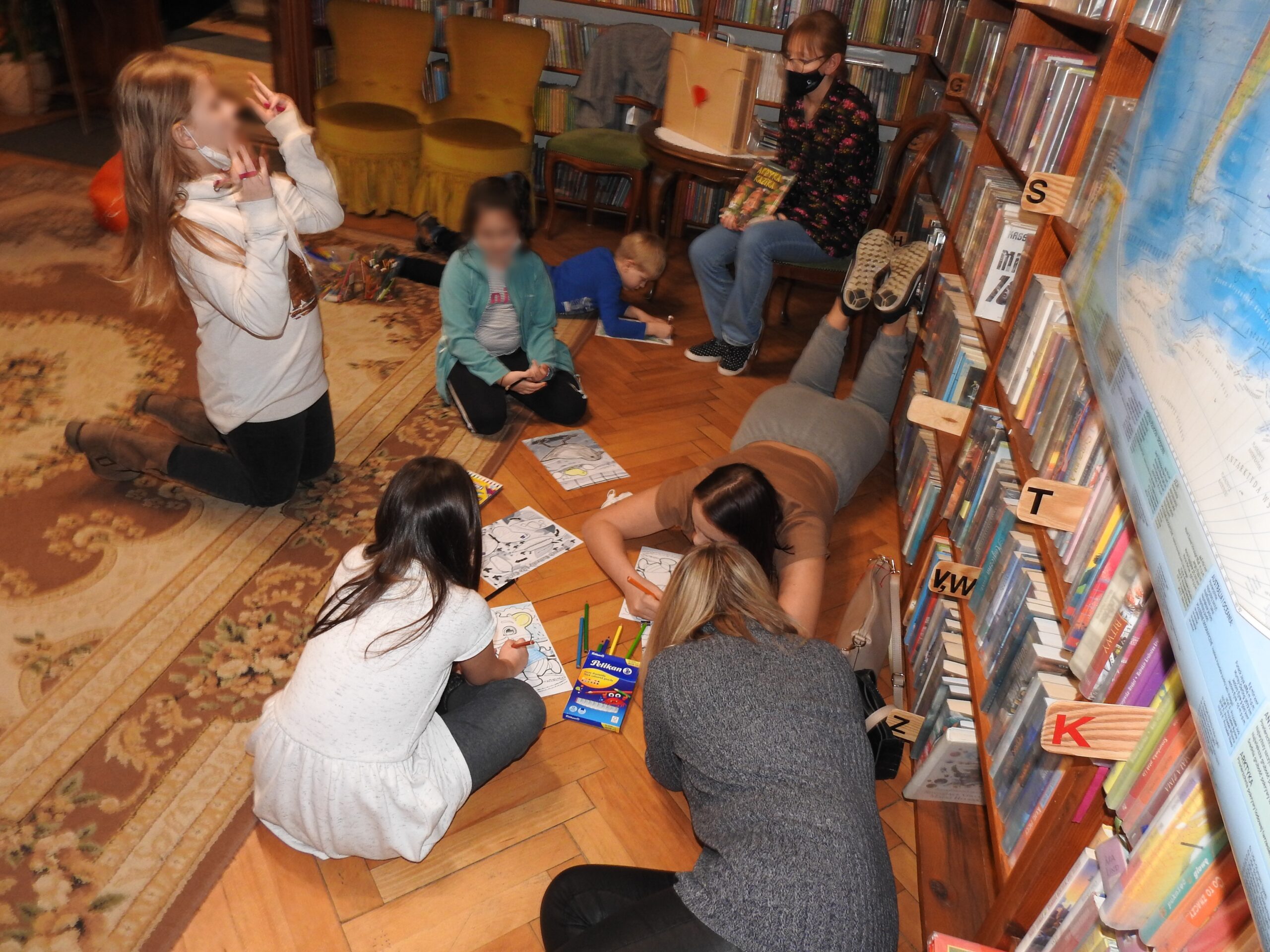 Cztery osoby w pomieszczeniu z ksiązkami. Dwie leżą na podłodze, dwie siedzą, przed nimi rysunki i kredki. Pita osoba siedzi na krześle.