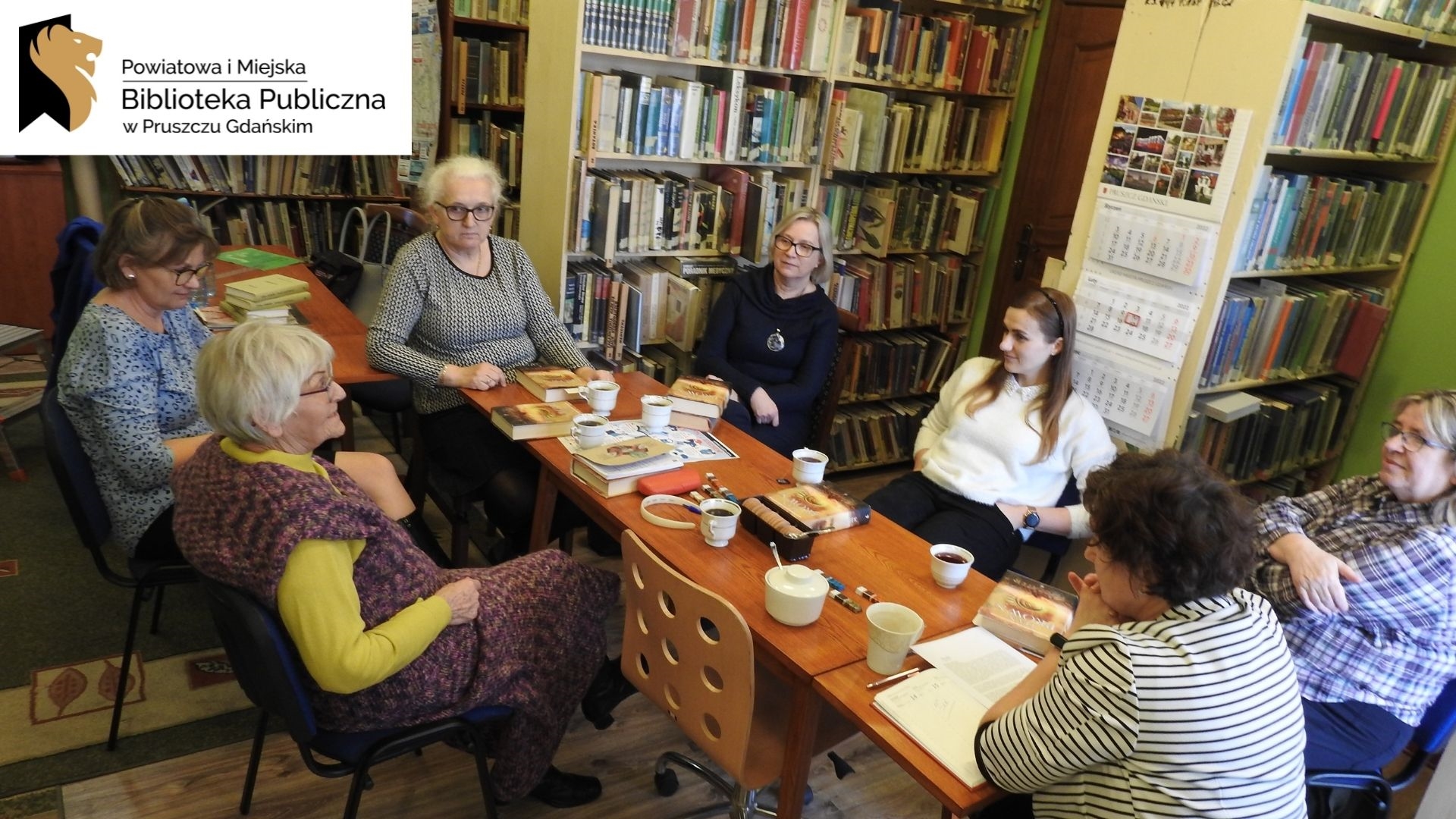 Sześć osób siedzi przy stolew pomieszczeniu z książkami. Logotyp oraz napis Powiatowa i Miejska Biblioteka Publiczna w Pruszczu Gdańskim.