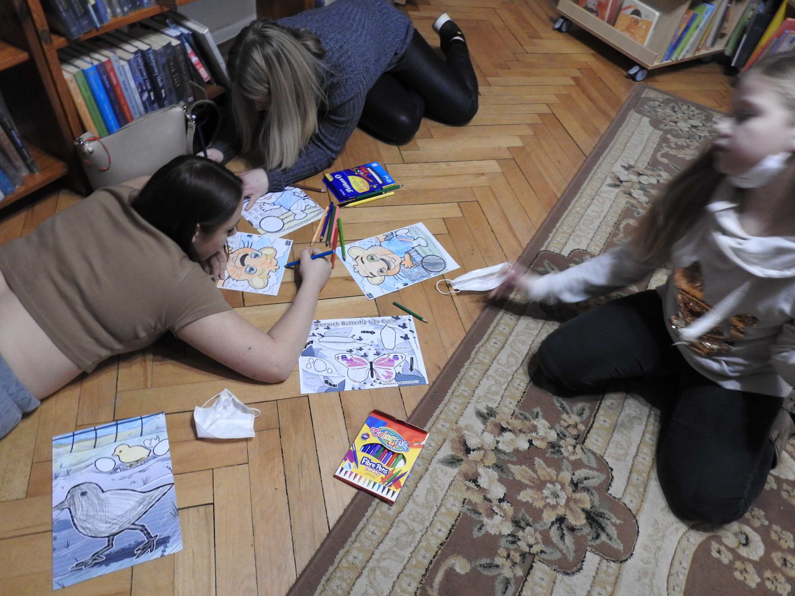 Cztery osoby w pomieszczeniu z ksiązkami. Dwie leżą na podłodze, dwie siedzą, przed nimi rysunki i kredki.