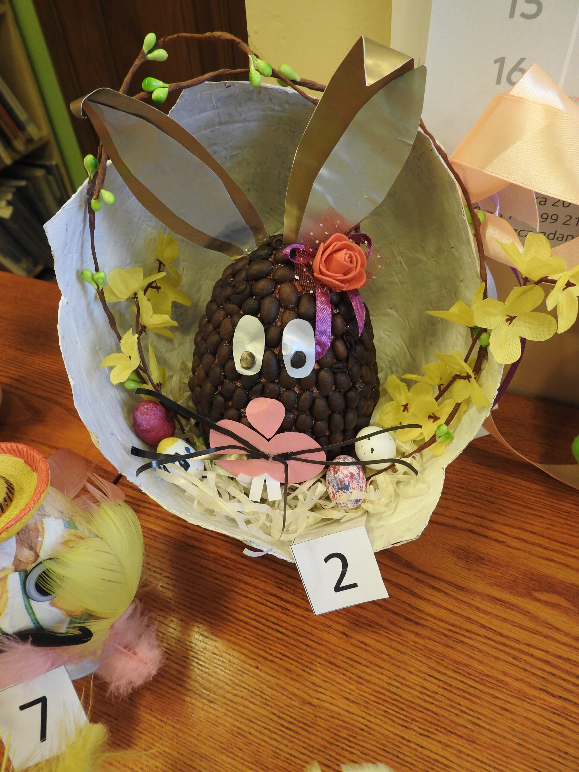Pisanka w formie zajączka: jajo oklejone ziarenkami kawy, doklejone papierowe uszy,. Całośc umieszczona w otwartm kole, ozodobiona sztucznymi kwiatkami, jajeczkami.