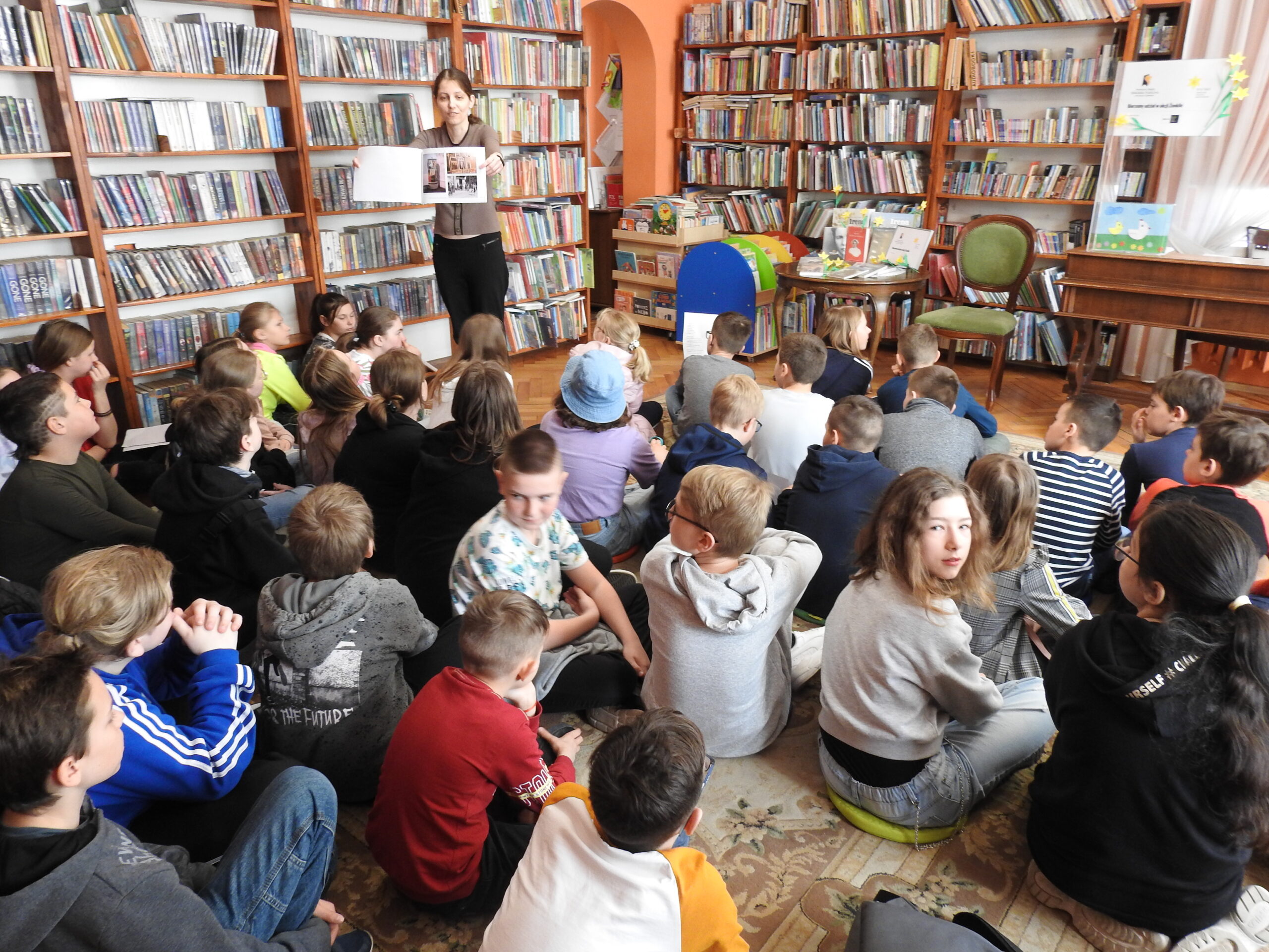 Grupa osób siedzi na dywanie w pomieszczeniu z książkami. Przed nimi stoi inna osoba, która trzyma otwartą książkę.