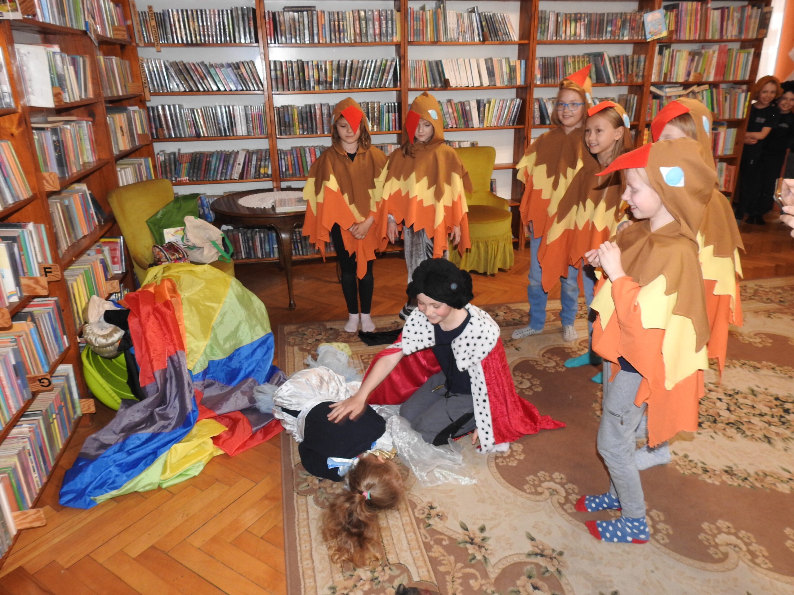 Jedna osoba leży na dywanie. Przy niej klęczy chłopiec w stroju księcia. Nad nimi stoi 6 osób w kostiumach ptaków. Po lewej stronie leżą kolorowe materiały. W tle stół, dwa fotele oraz regały z książkami.