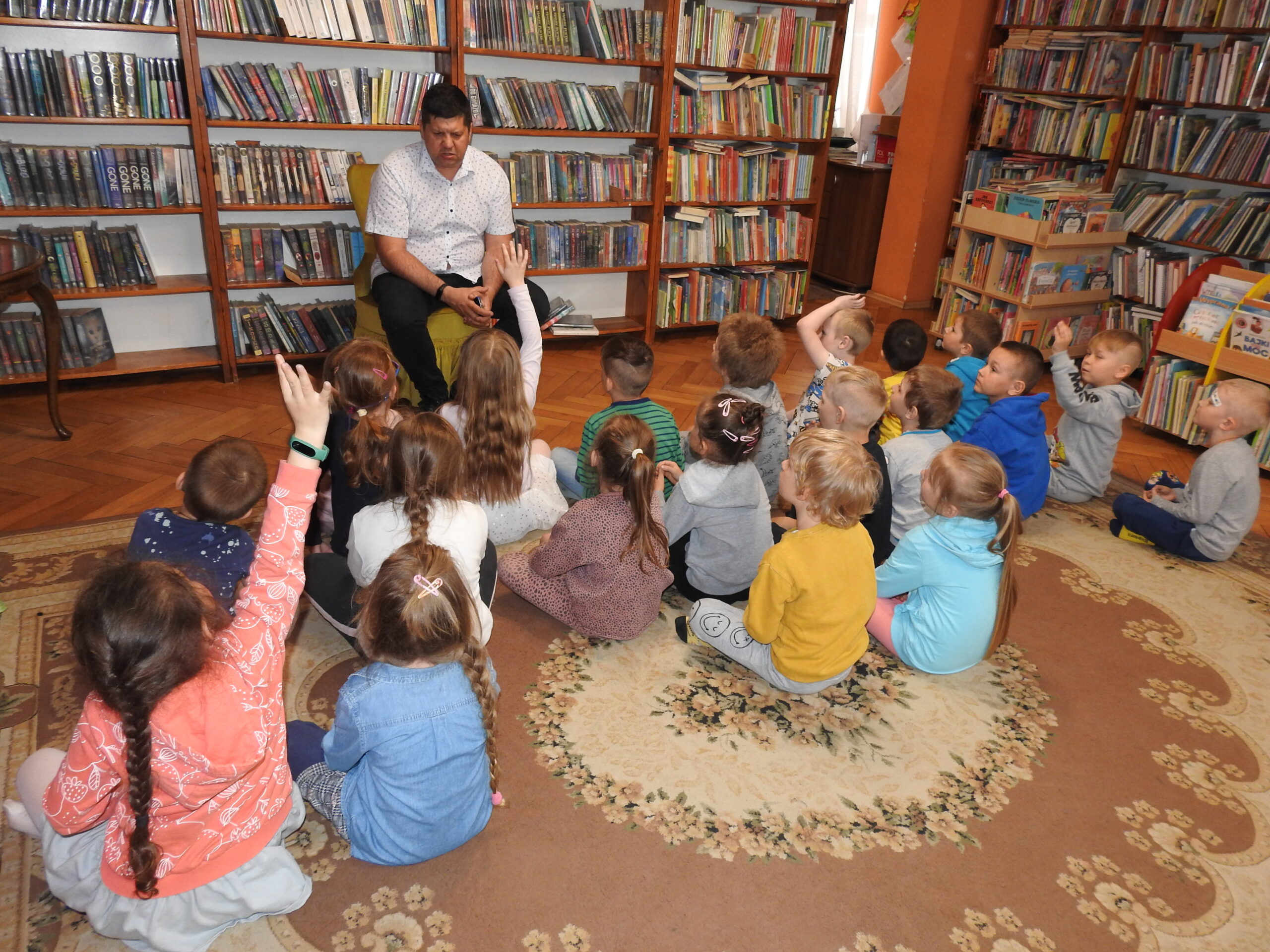 Grupa dzieci siedzi na dywanie. Jest skierowana w stronę mężczyzny, który siedzi na fotelu. Czwórka dzieci trzyma rękę w górze. W tle regały z książkami.