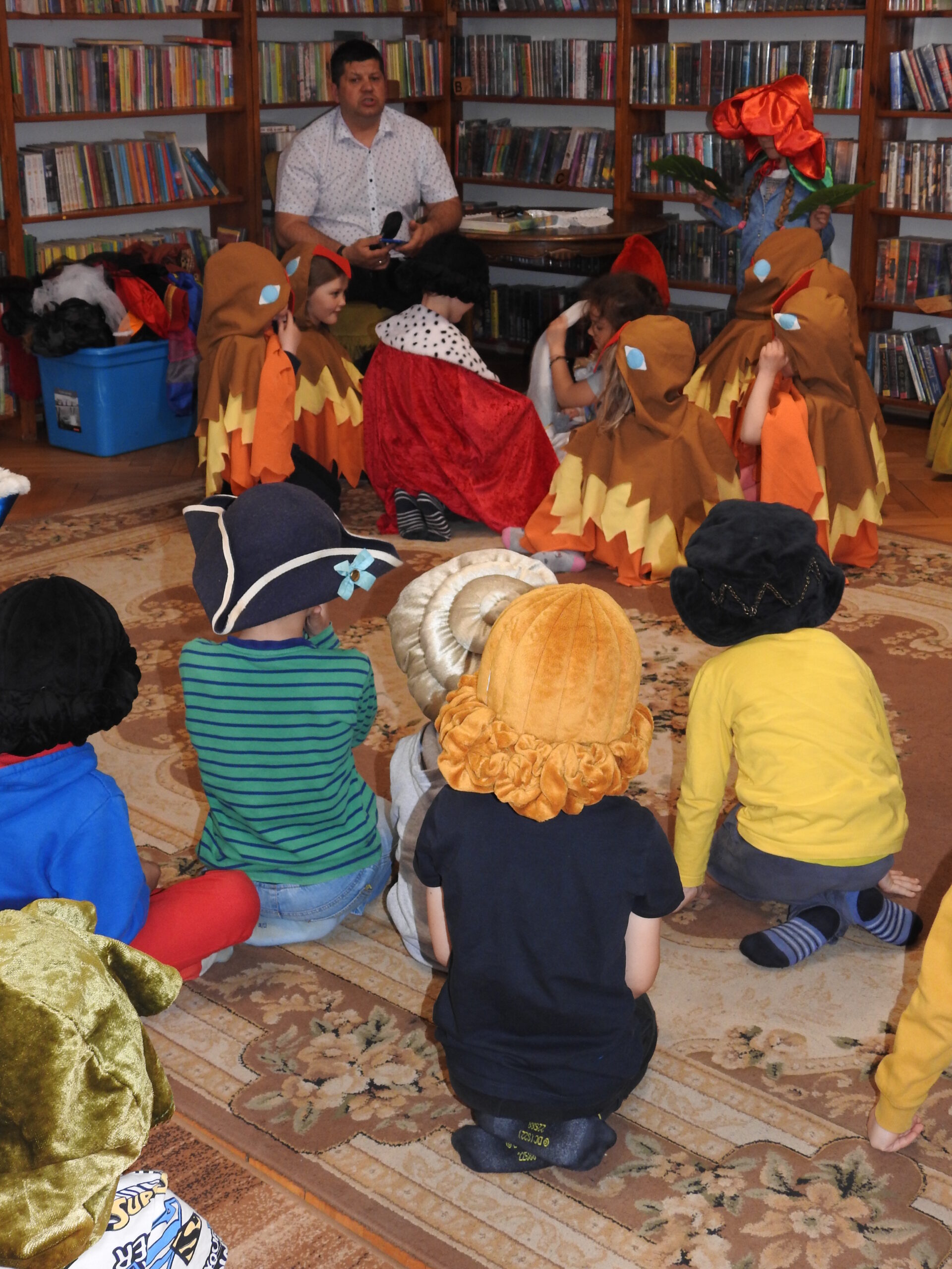 Grupa dzieci ubrana w kostiumy siedzi na dywanie. Jest skierowana w stronę mężczyzny siedzącego na fotelu. W tle regały z książkami.