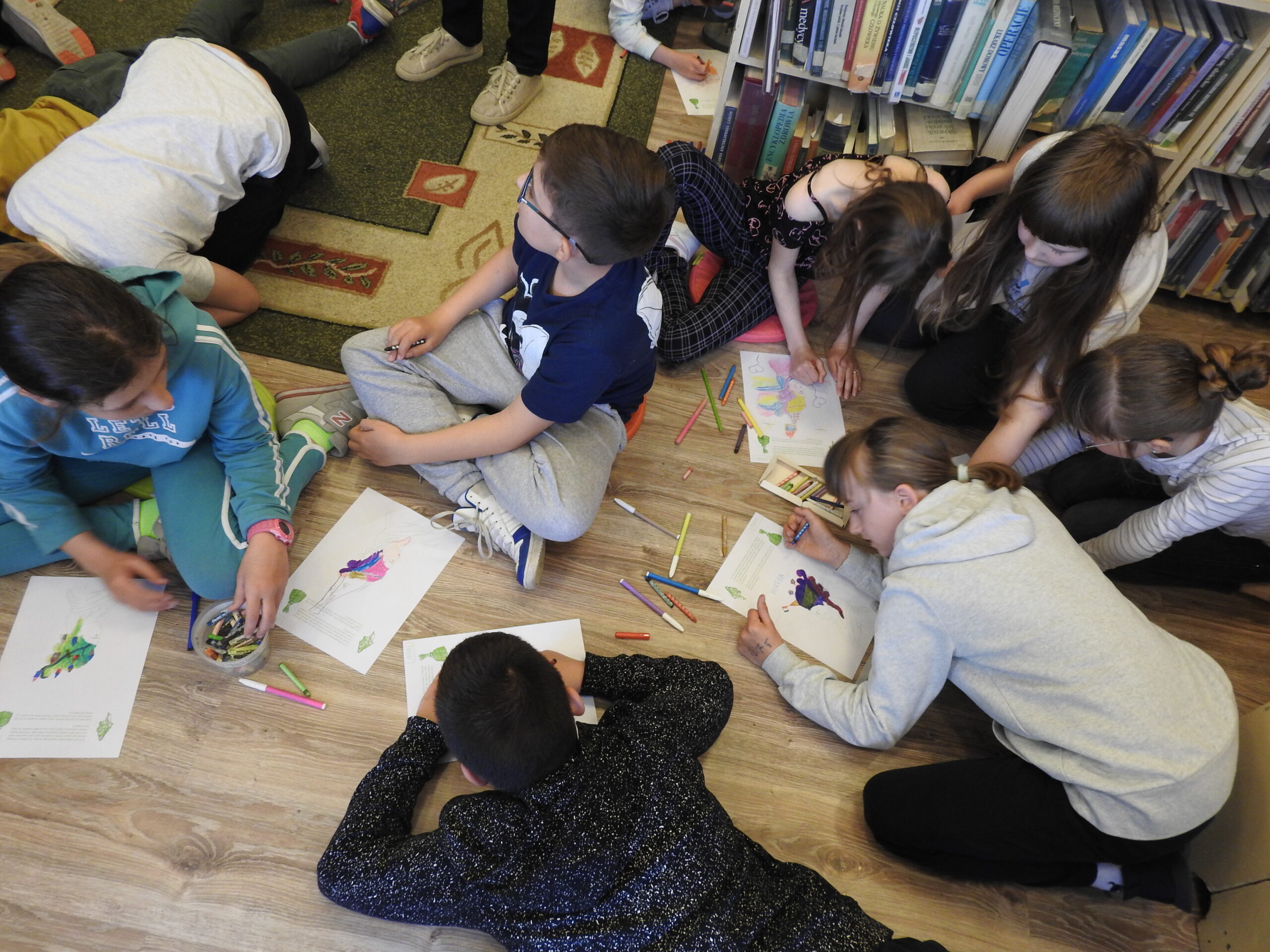 Grupa dzieci siedzi na podłodze i koloruje obrazki. Wokół nich porozkładane są mazaki i kredki. 