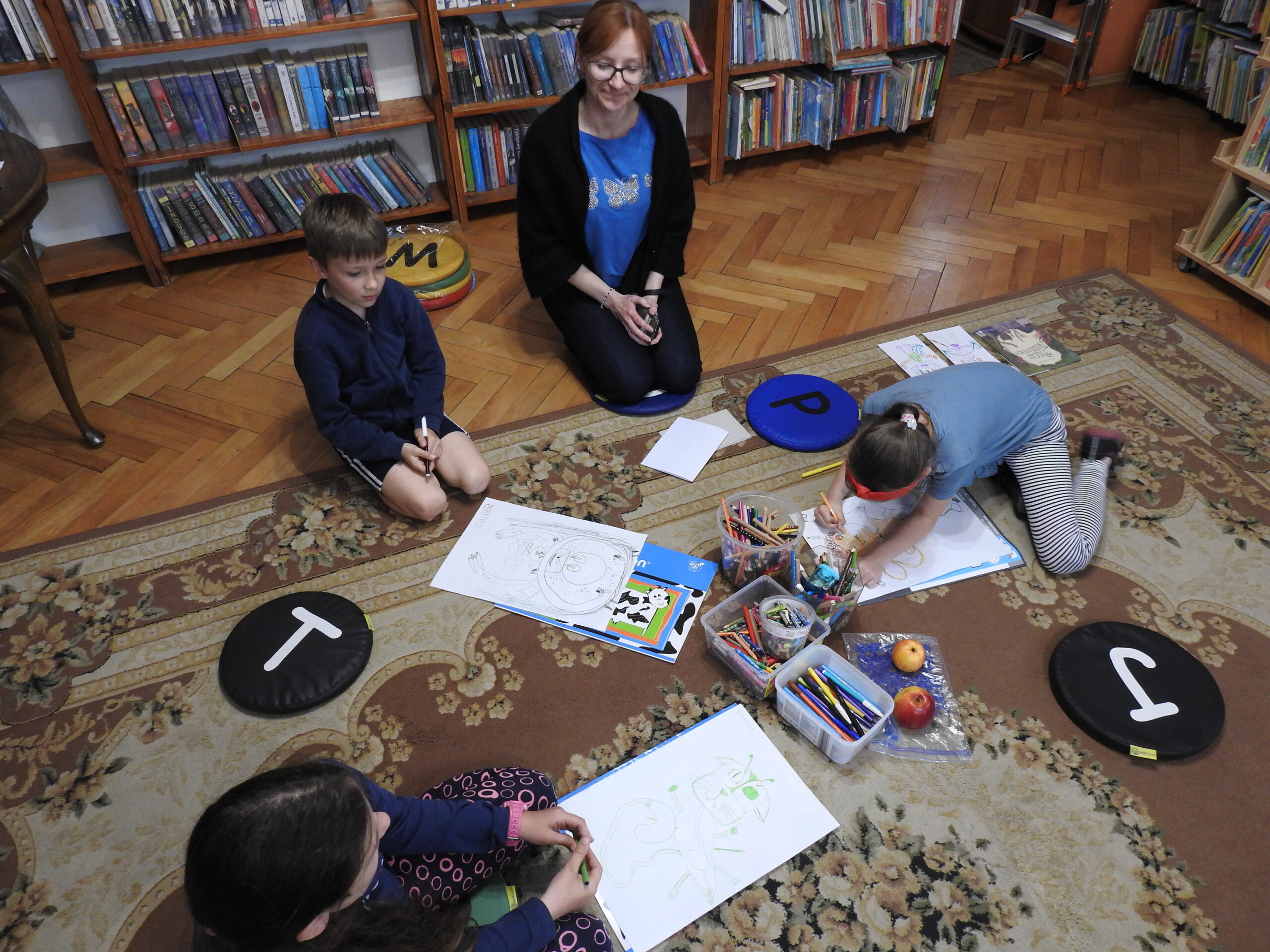 Troje dzieci oraz jedna osoba dorosła siedzą na dywanie i podłodze. Przed dziećmi znajdują się duże kartki, na których namalowane są potwory. Jedno dziecko pochyla się nad kartką i jeszcze coś dorysowuje. W tle regały z książkami.