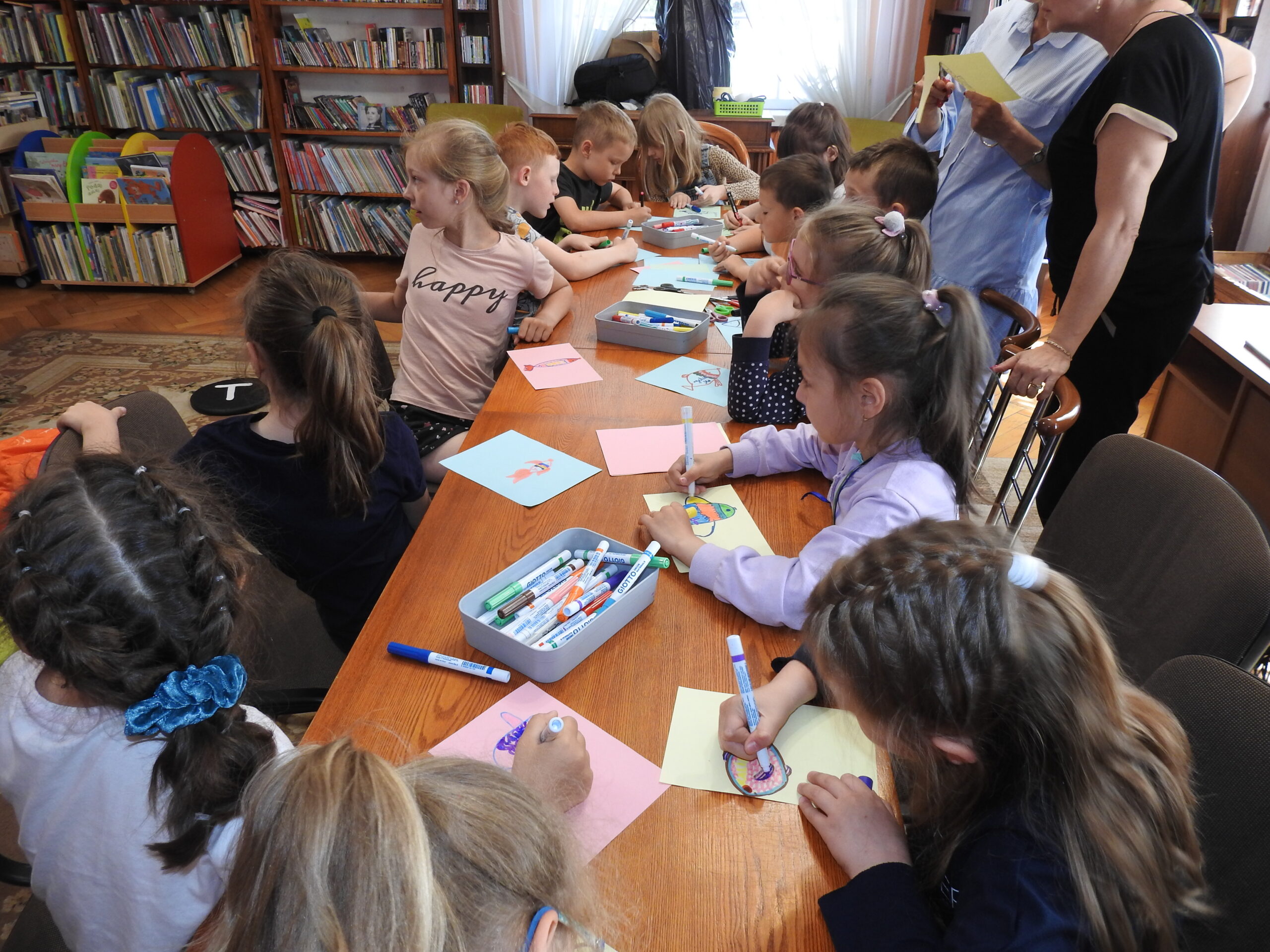 Grupa dzieci siedzi wokół stołu. Przed nimi, na stole, leżą kolorowe kartki oraz mazaki. Część dzieci maluje na kartkach flamastrami, a część jest zwrócona w lewą stroną, w kierunku czegoś, co znajduje się poza zdjęciem. Dzieci malują ryby.