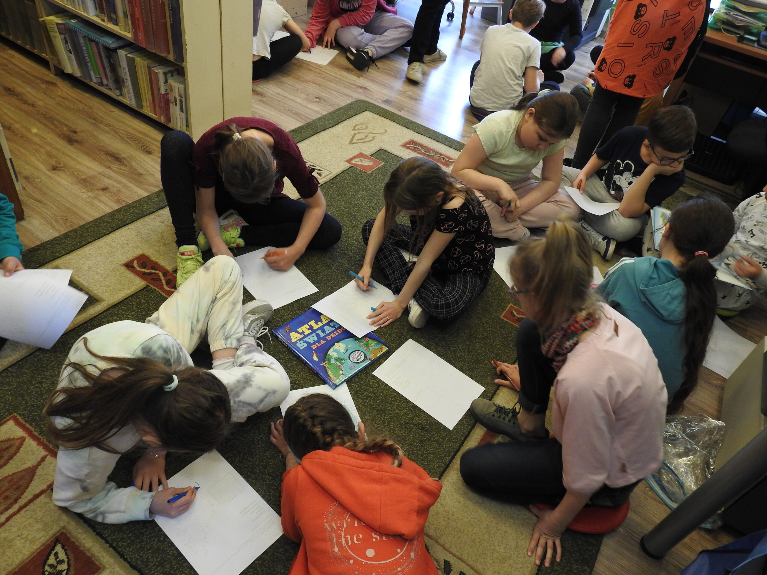 Grupa dzieci siedzi na dywanie. Każdy ma jedną kartkę A4. Część osób na niej pisze. Pomiędzy dziećmi leży Atlas świata dla dzieci.