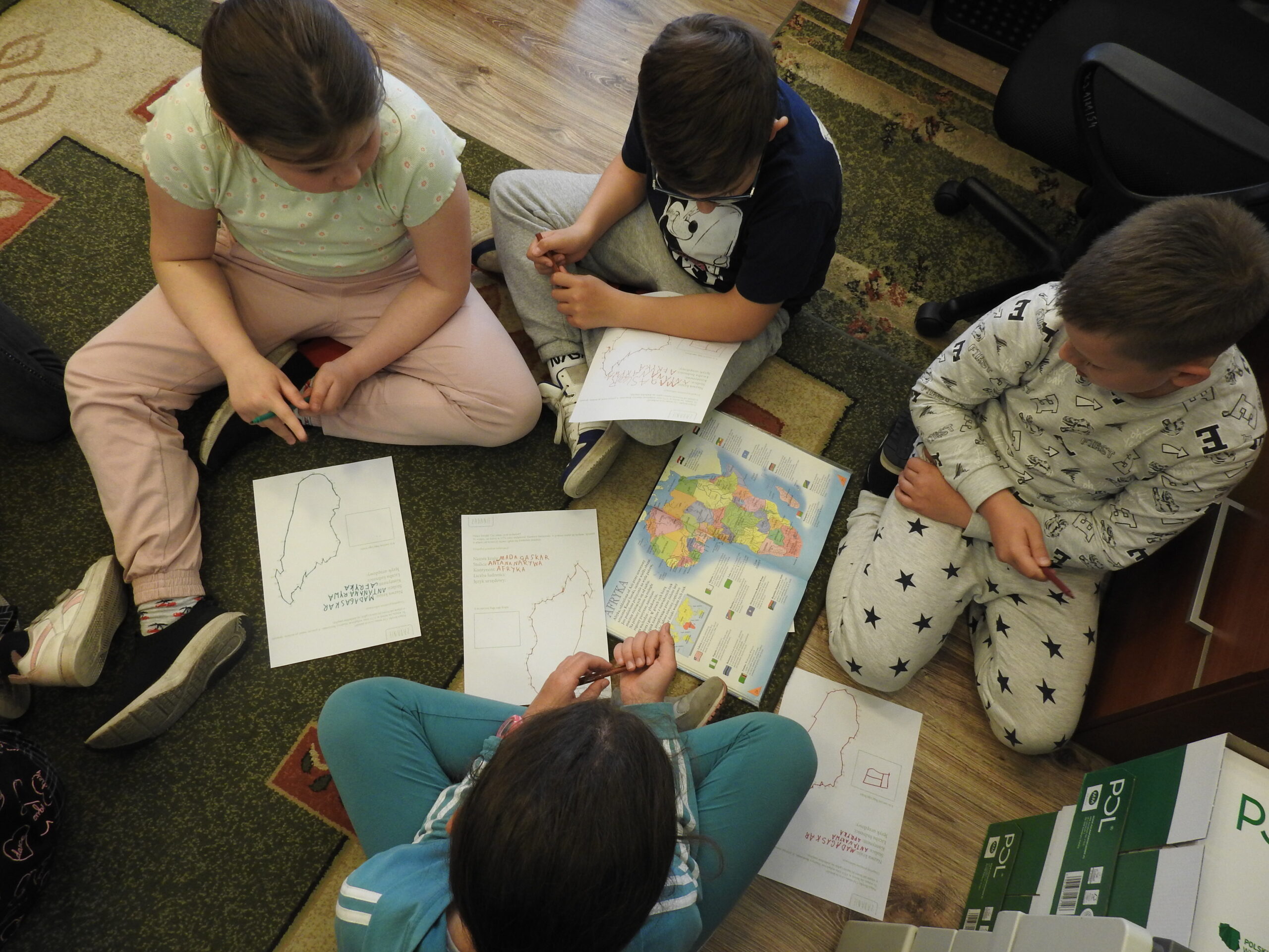 Czworo dzieci siedzi na dywanie wokół otwartego atlasu z mapą Afryki i czterech kartek A4. Na kartkach narysowane są kontury państwa. .