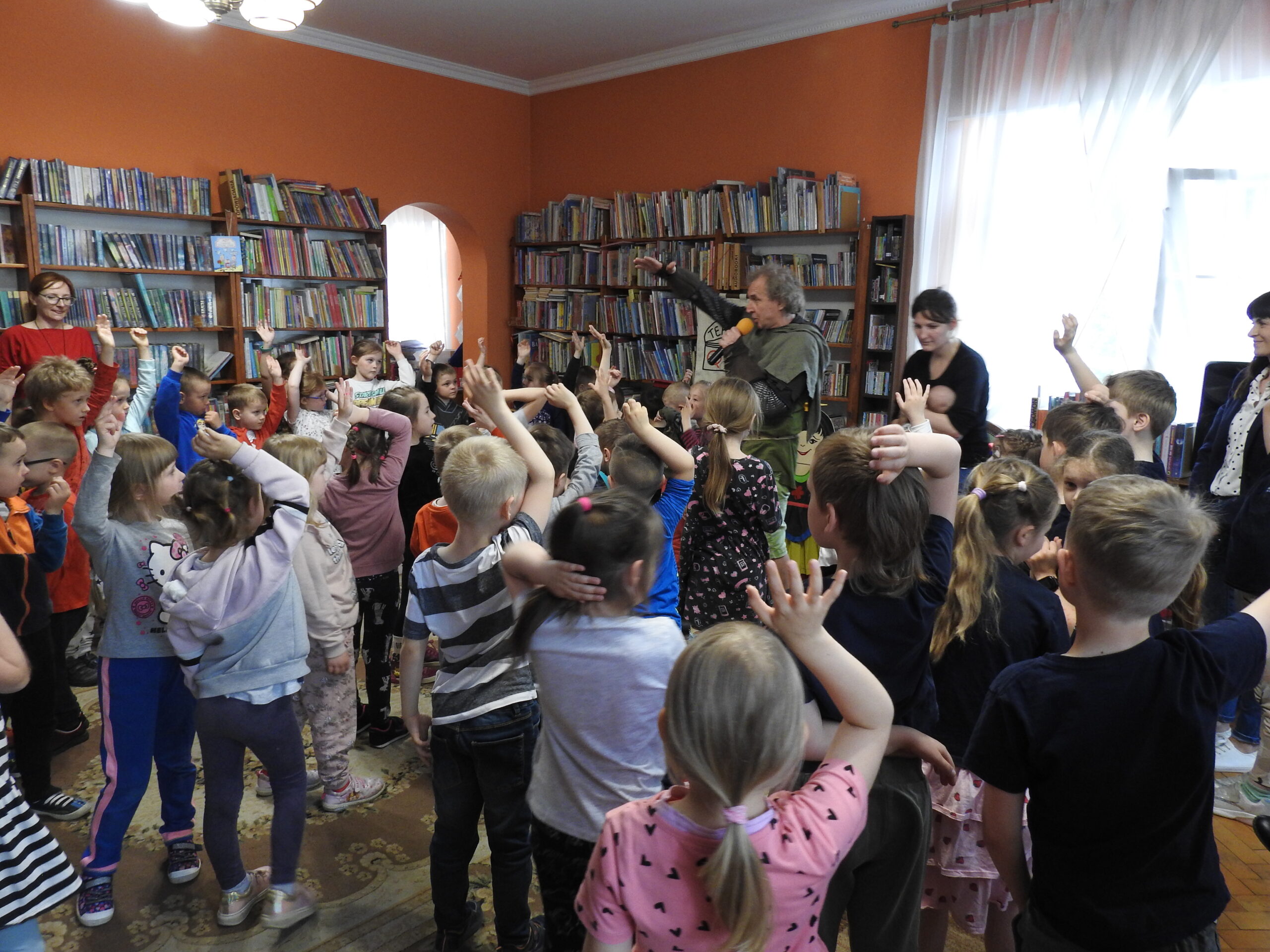 Duża grupa dzieci stoi zwrócona w stronę mężczyzny ubranego w zielono-brązowy kostium i trzymającego mikrofon. Wszyscy trzymają jedną rękę w górze. W tle regały z książkami. 