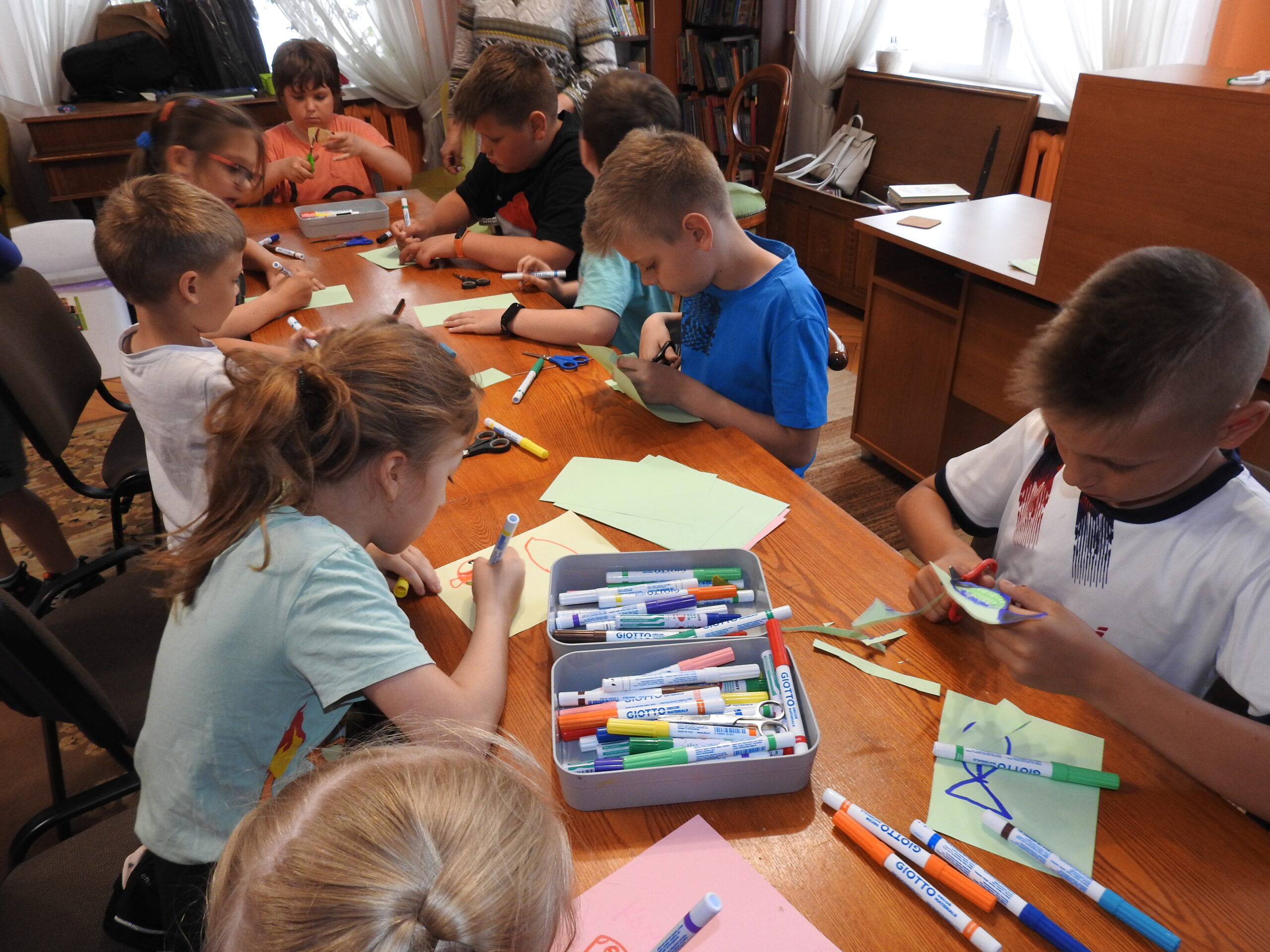 Grupa dzieci siedzi przy stole. Przed nimi, leżą kolorowe kartki oraz flamastry. Część dzieci maluje na kartkach, a część wycina.
