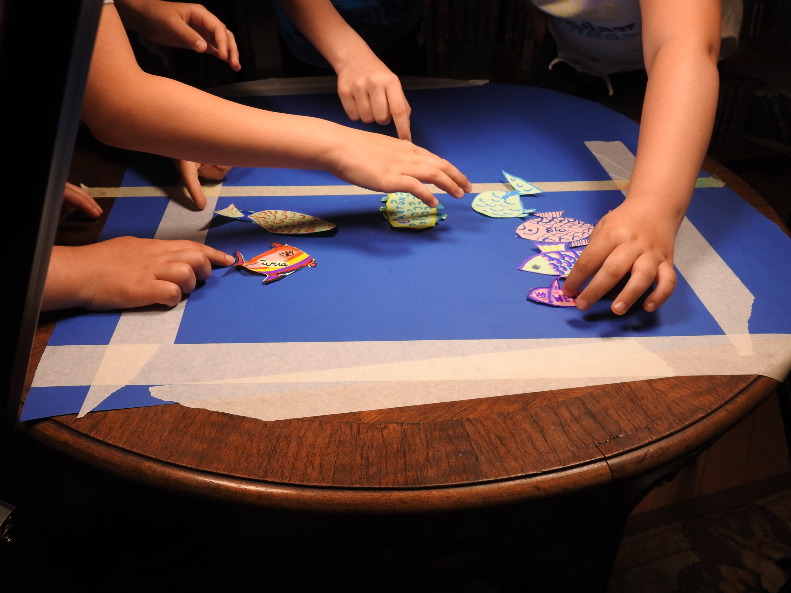 Na stoliku leży niebieski arkusz papieru z wyznaczonym białą taśmą prostokątem. Ręce układają kolorowe rybki na tym prostokącie.