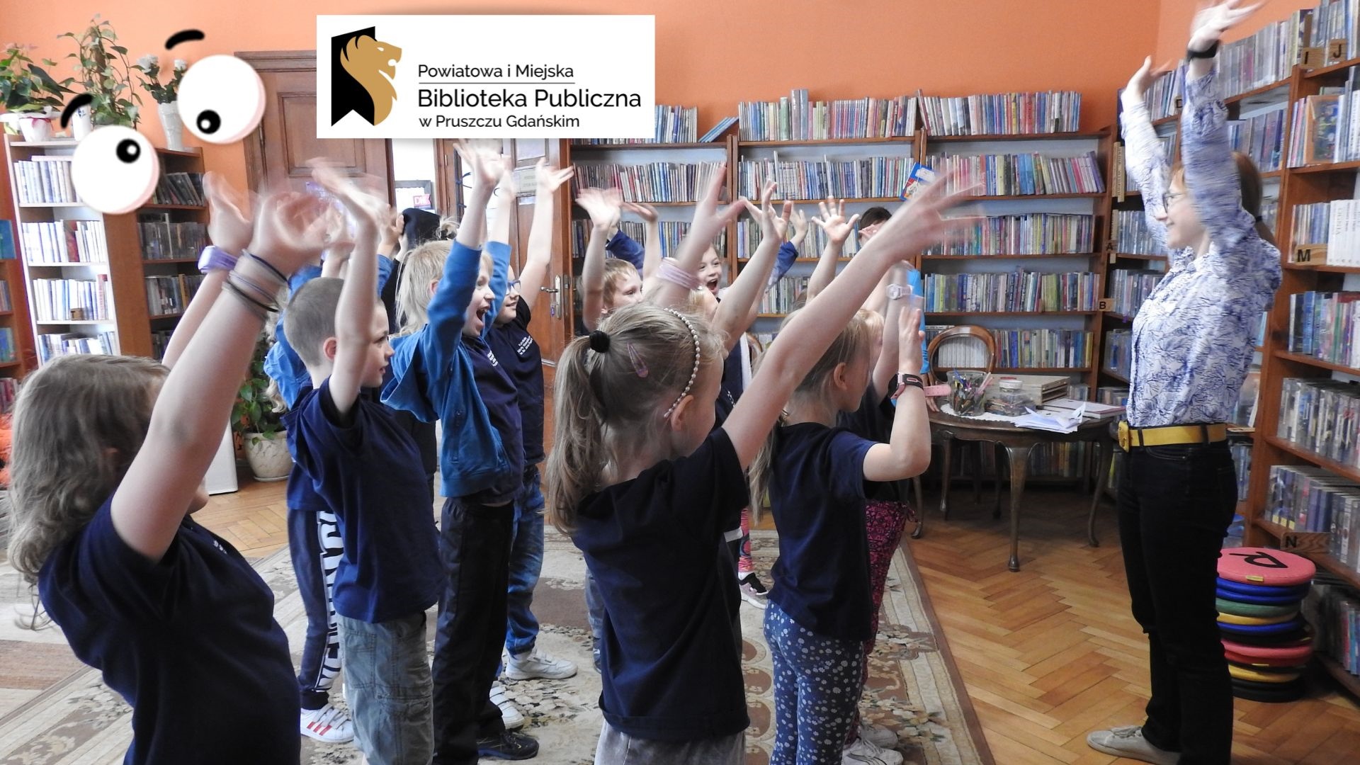 Grupa dzieci stoi naprzeciwko osoby dorosłej. Wszyscy trzymają ręce w górze. W tle regały z książkami. W lewym górnym rogu logotyp oraz napis: Powiatowa i Miejska Biblioteka Publiczna w Pruszczu Gdańskim.