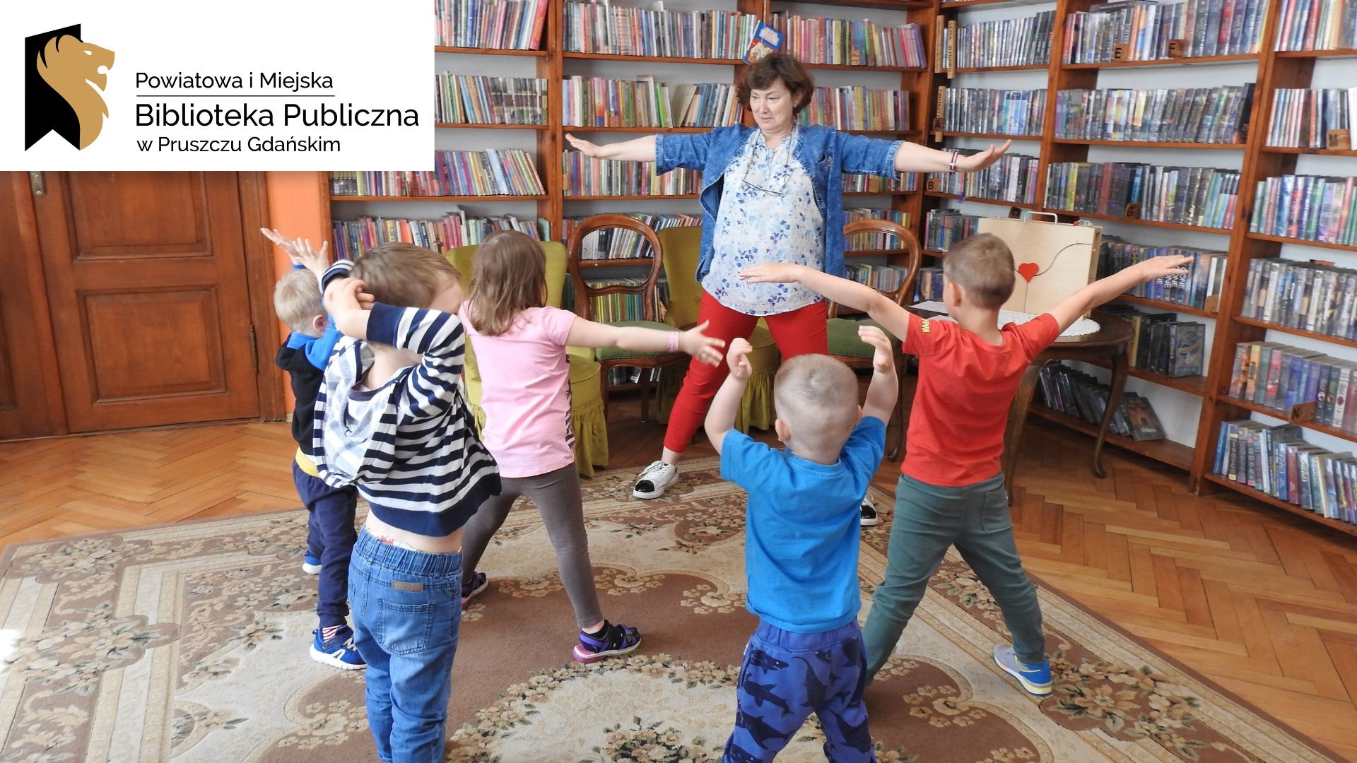 Kilkoro dzieci stoi naprzeciwko osoby dorosłej. Wszyscy mają szeroko rozłożone ręce. W tle regały z książkami.Logotyp oraz napis: Powiatowa i Miejska Biblioteka Publiczna w Pruszczu Gdańskim.