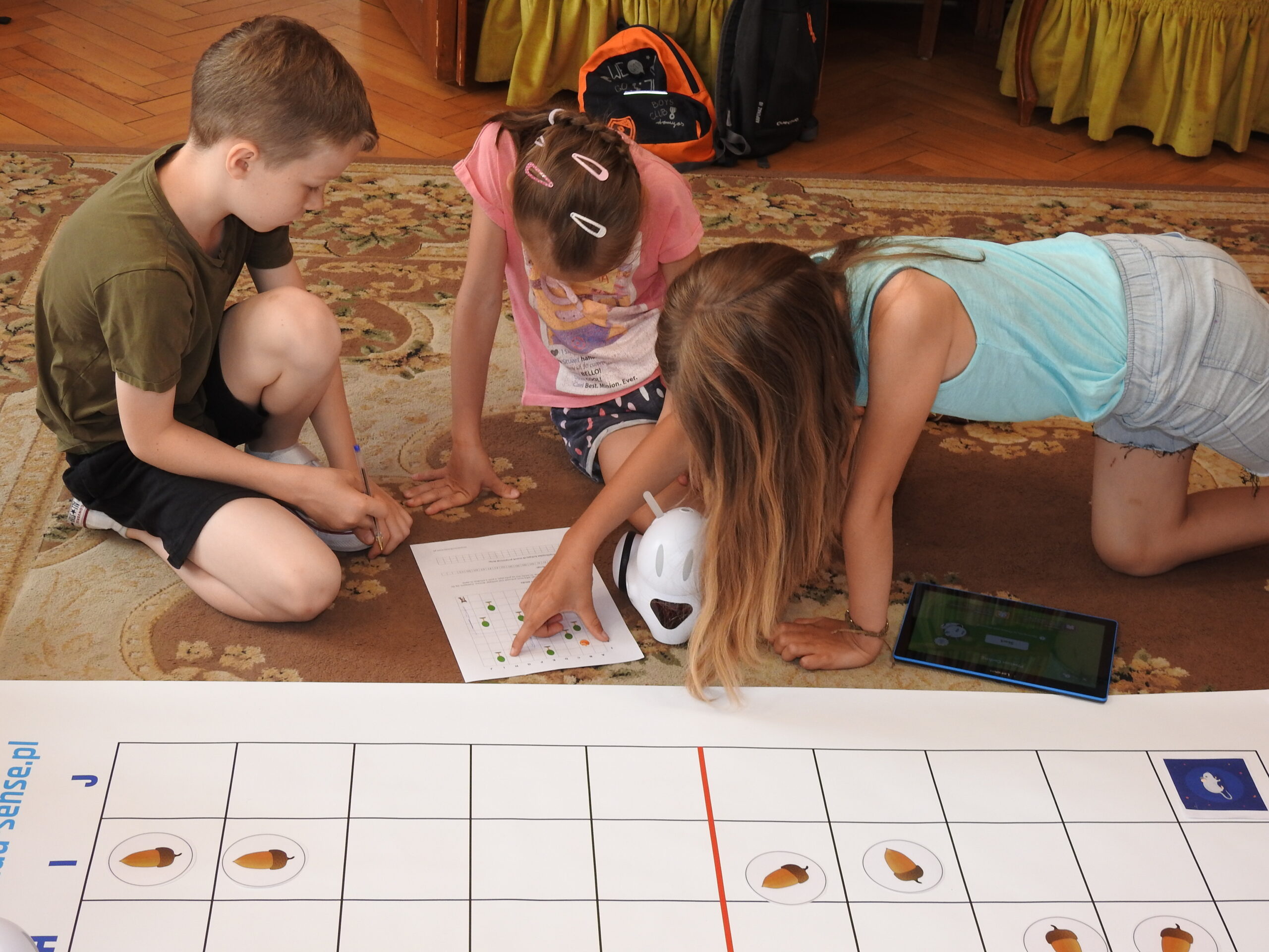 Troje dzieci siedzi lub klęczy na dywanie i jest pochylonych nad kartką A4 z nadrukowaną instrukcją do kodowania. Na kartce widać planszę podzieloną na kwadraty. W kilku kwadratach znajdują się zielone i żółte kółeczka. Jedno dziecko trzyma rękę na kartce wskazując palcami określone kwadraty. Obok dzieci znajduje się biały robot do kodowania oraz tablet. Widać też fragment maty do kodowania.