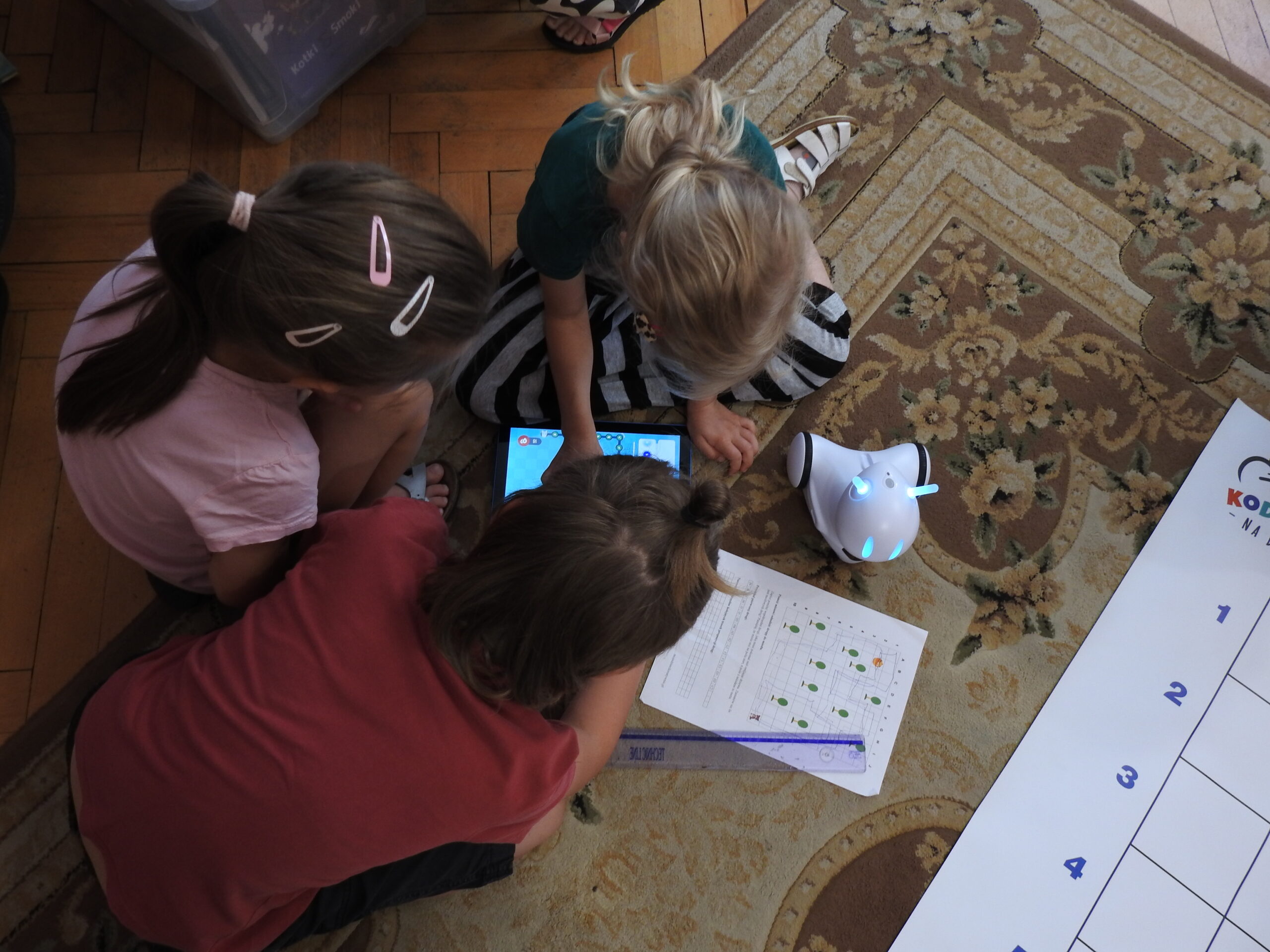 Troje dzieci siedzi na dywanie i jest pochylonych nad włączonym tabletem. Obok nich znajduje się robot do kodowania oraz kartka A4 z nadrukowaną instrukcją do kodowania. Na kartce widać planszę podzieloną na kwadraty. W kilku kwadratach znajdują się zielone i żółte kółeczka. 