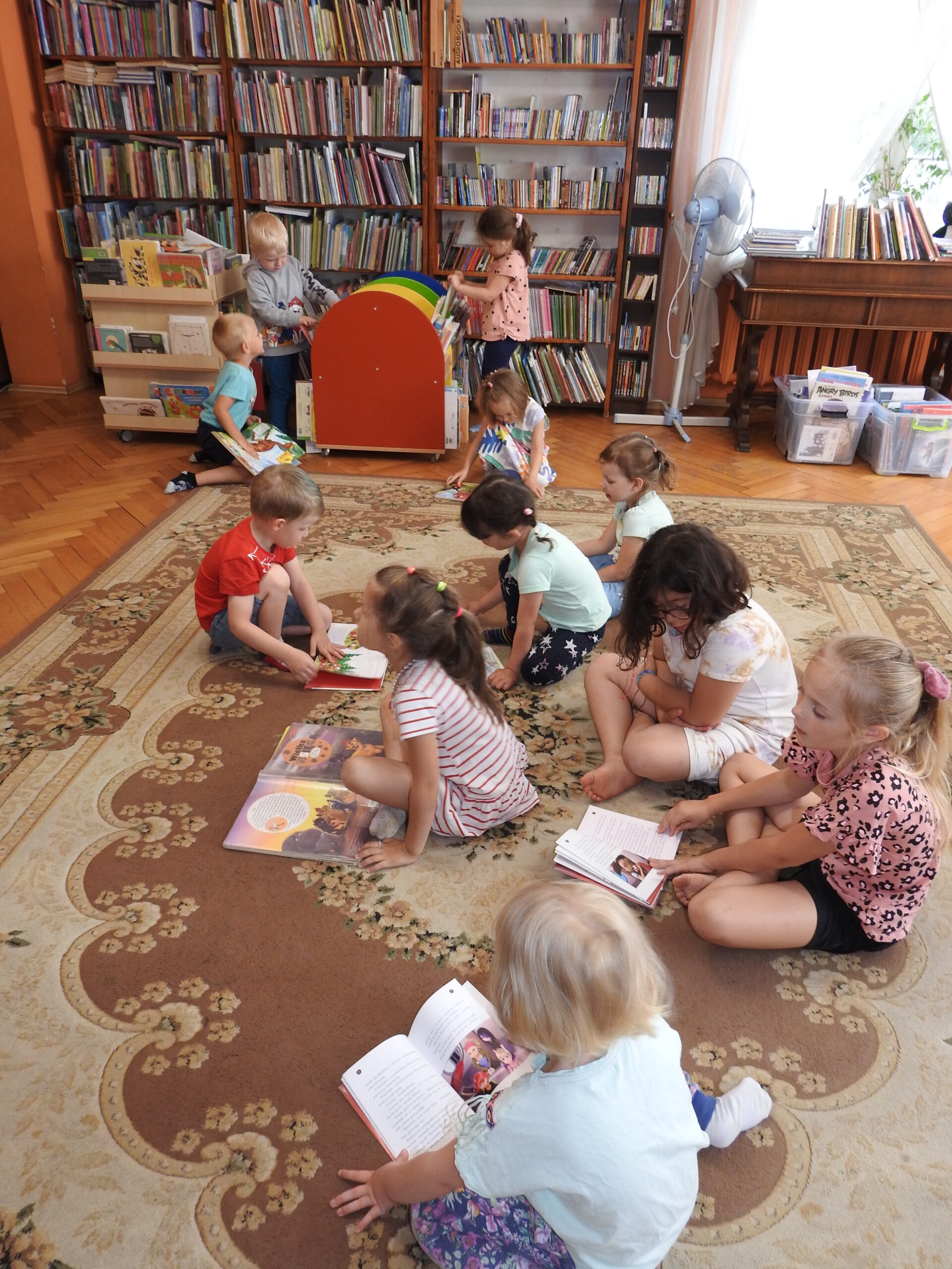 Grupa dzieci siedzi na dywanie. Przed nimi znajdują się otwarte książki. Troje dzieci ogląda książki na małym regale na kółkach. W tle tradycyjne regały z książkami.