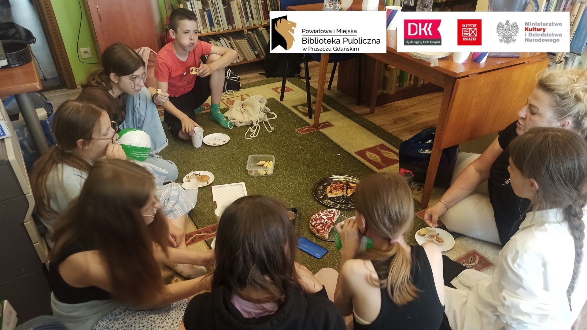 Dzieci z Dyskusyjnego Klubu Książki wraz z moderatorką siedzą na dywanie w okręgu. Na dywanie postawione są papierowe talerzyki, kubeczki, ciasta, rogaliki, babeczki.